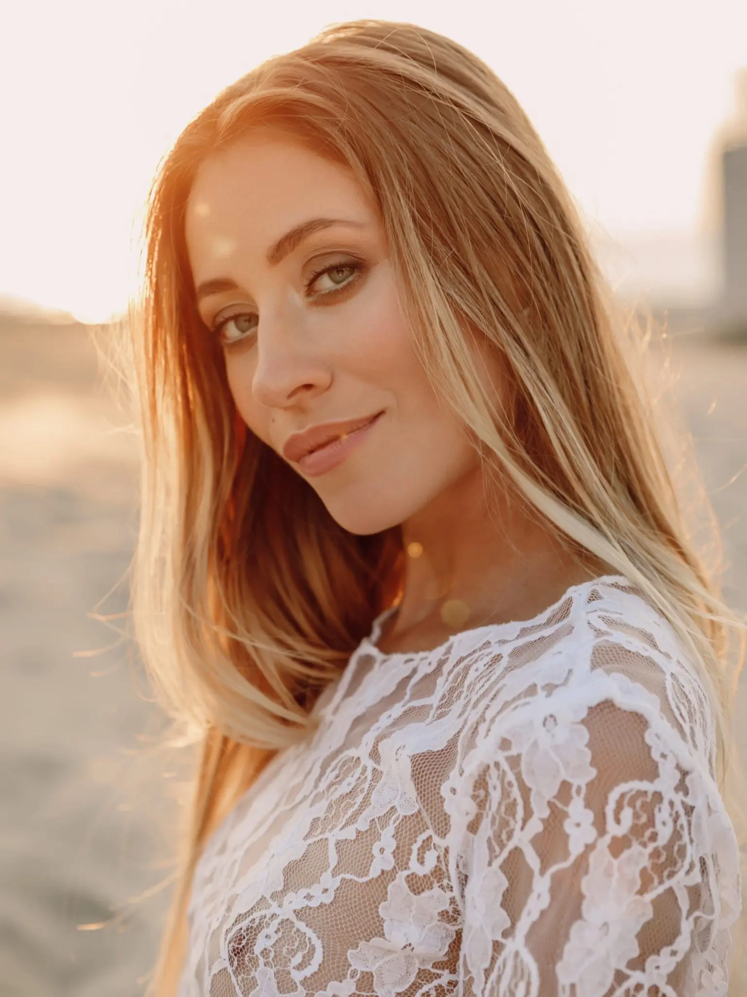 A woman with long, light brown hair and a lace white top stands outdoors at sunset, looking at the camera with a slight smile—radiant and confident after Juvederm treatment.