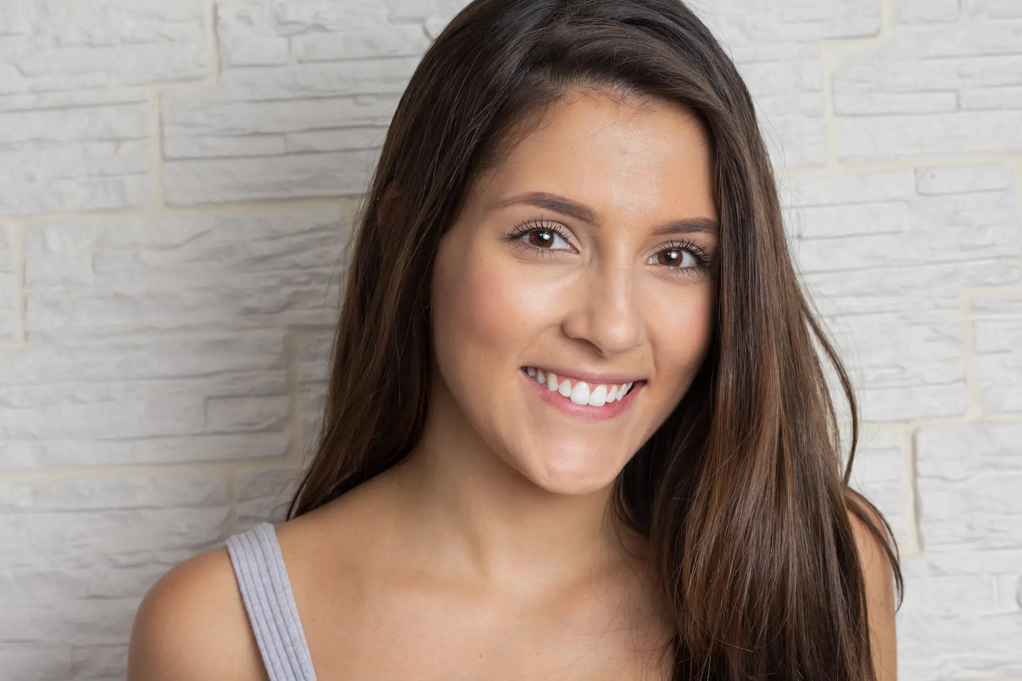 Smiling, confident young woman with long brown hair in gray tank top after Skin Resurfacing at Refresh MedSpa.