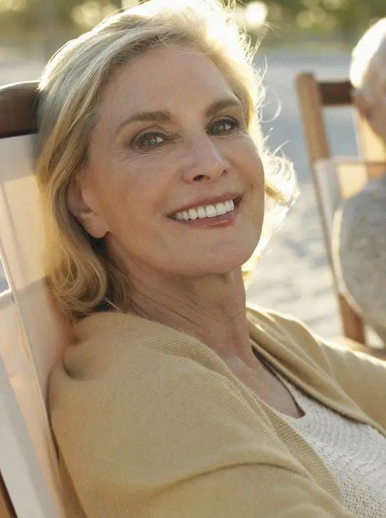 A woman with blonde hair sits outdoors on a lounge chair, smiling at the camera. She is wearing a beige sweater over a white top, enjoying her day after receiving Sculptra.