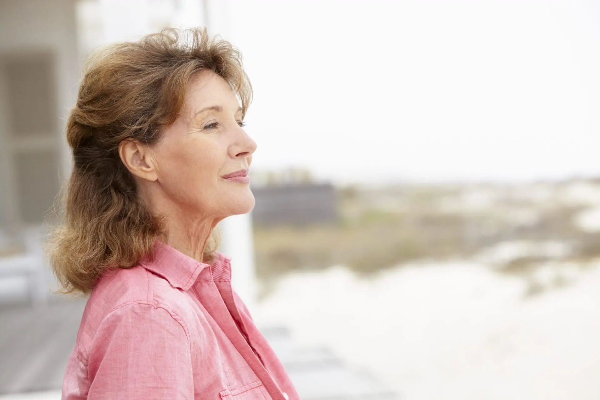 Middle-aged woman in a pink shirt stands outside, looking into the distance with a calm expression—an inspiring example of natural beauty enhanced by Radiesse.