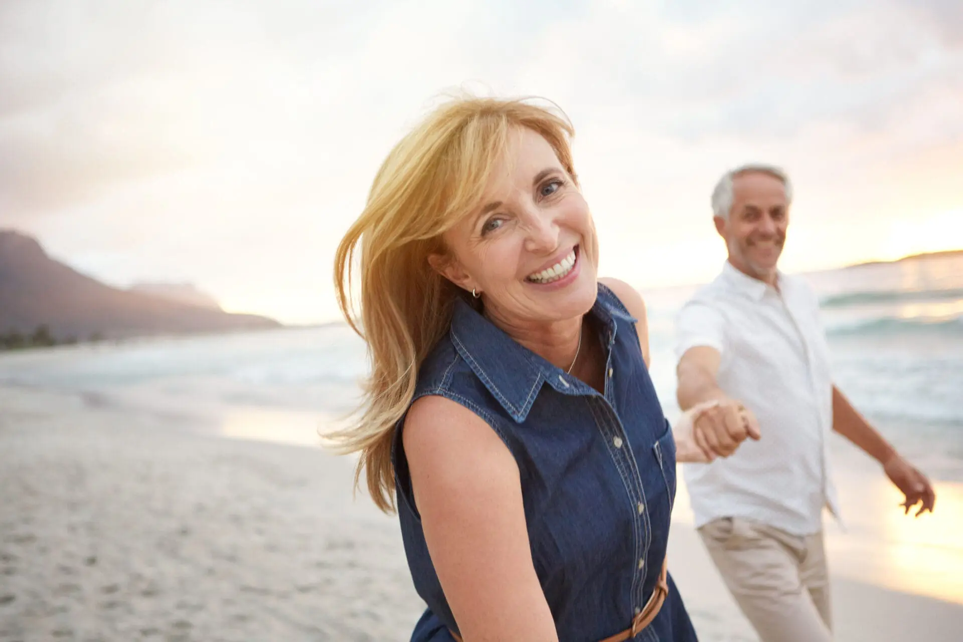 A woman smiles at the camera while holding hands with a man as they walk together on a sandy beach at sunset, enjoying their time after a Restylane treatment.