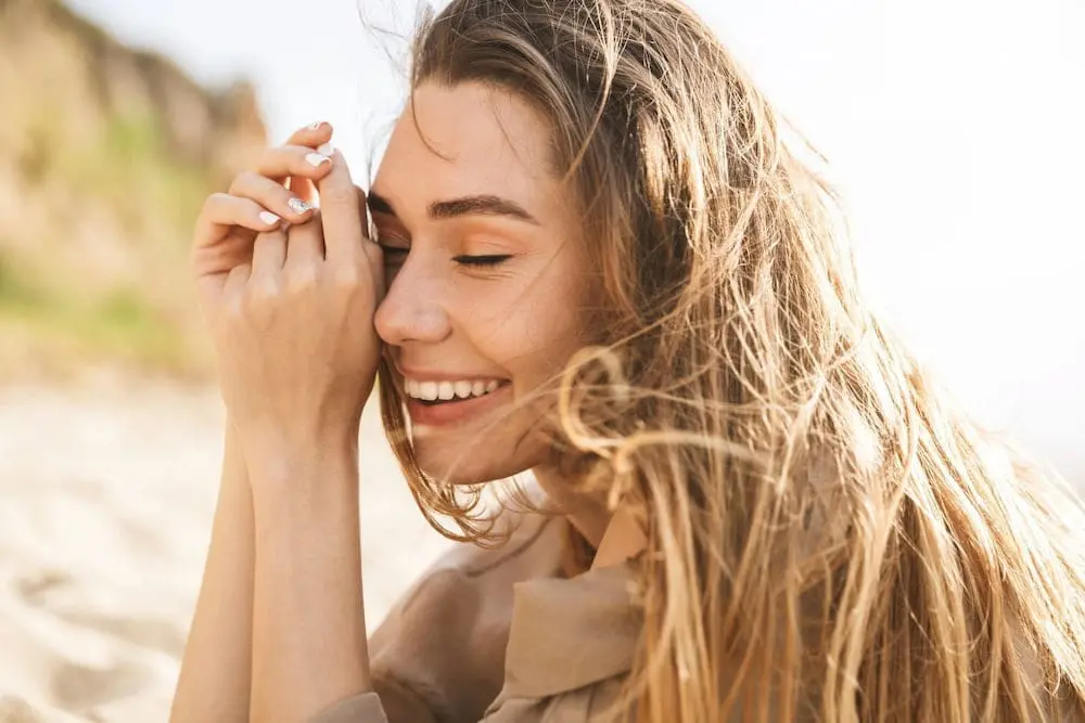 A woman with long hair smiles with eyes closed on a sunny beach, reflecting the calm Refresh MedSpa promotes.