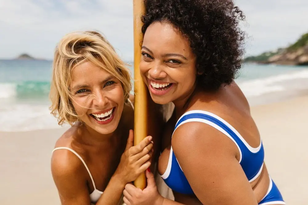 Two women in swimsuits smile confidently on a Cape Coral beach, possibly after injectables at Refresh MedSpa.