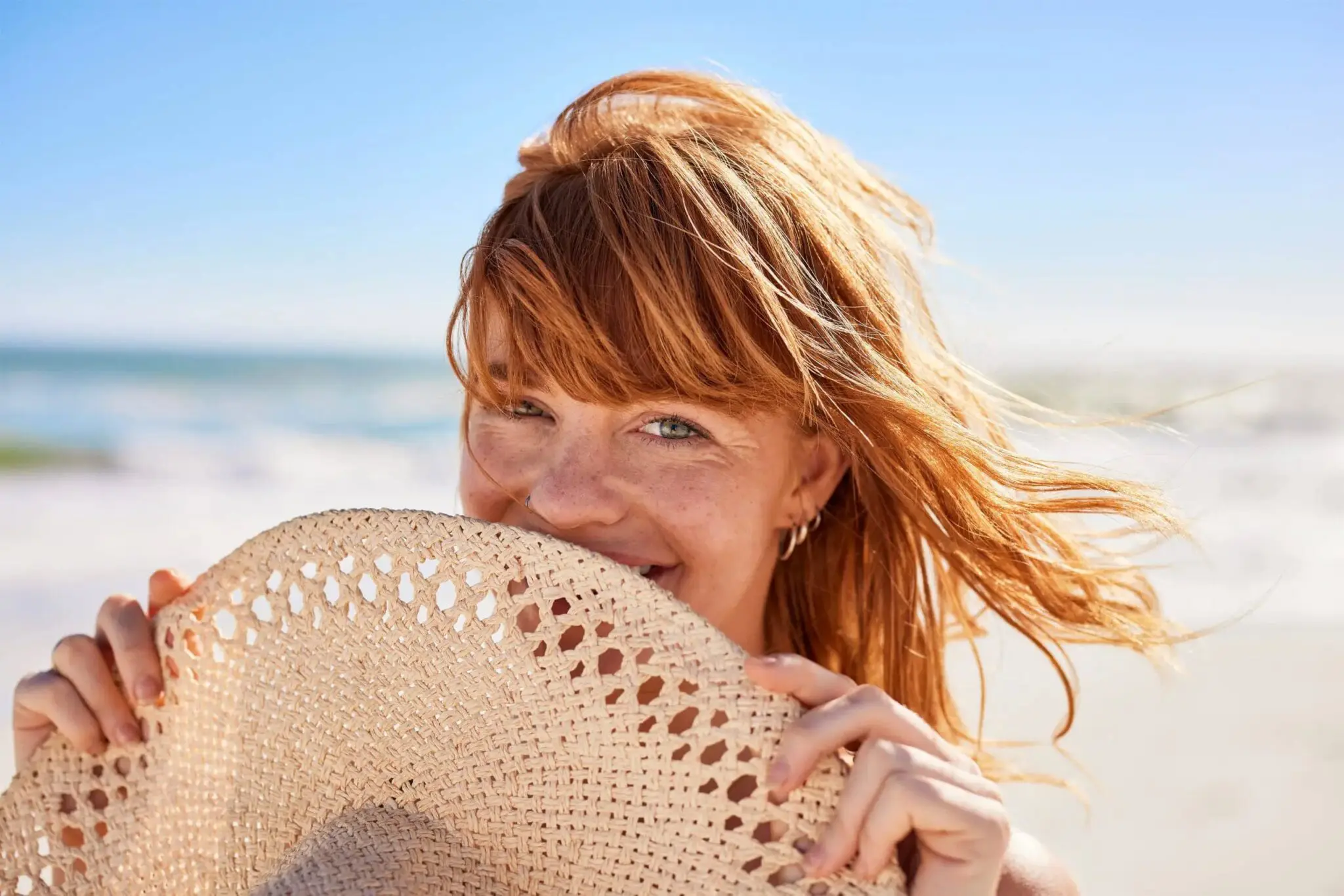 A person with red hair stands on a beach, holding a woven hat in front of their mouth and partially smiling, with the ocean and sky behind them—capturing the radiant confidence inspired by Refresh MedSpa, Best Medical Spa in Cape Coral, FL.