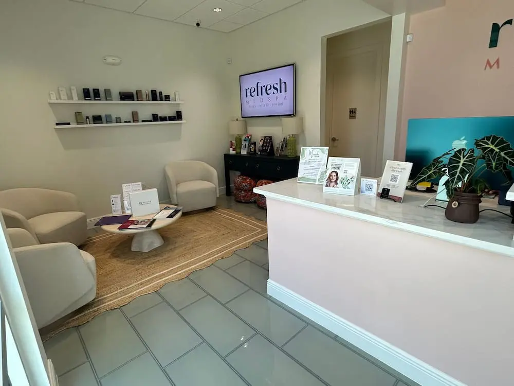 A modern spa reception area with a marble desk, informational brochures, wall-mounted shelves, chairs, and a rug. A digital screen displays the spa's logo at Refresh Medspa in Cape Coral, FL.