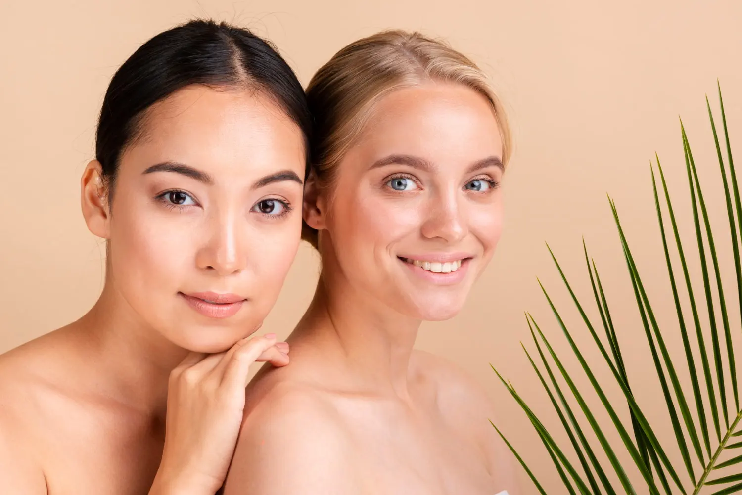 Two young women with bare shoulders pose closely together in front of a beige background with a green palm leaf in the foreground in Cape Coral FL