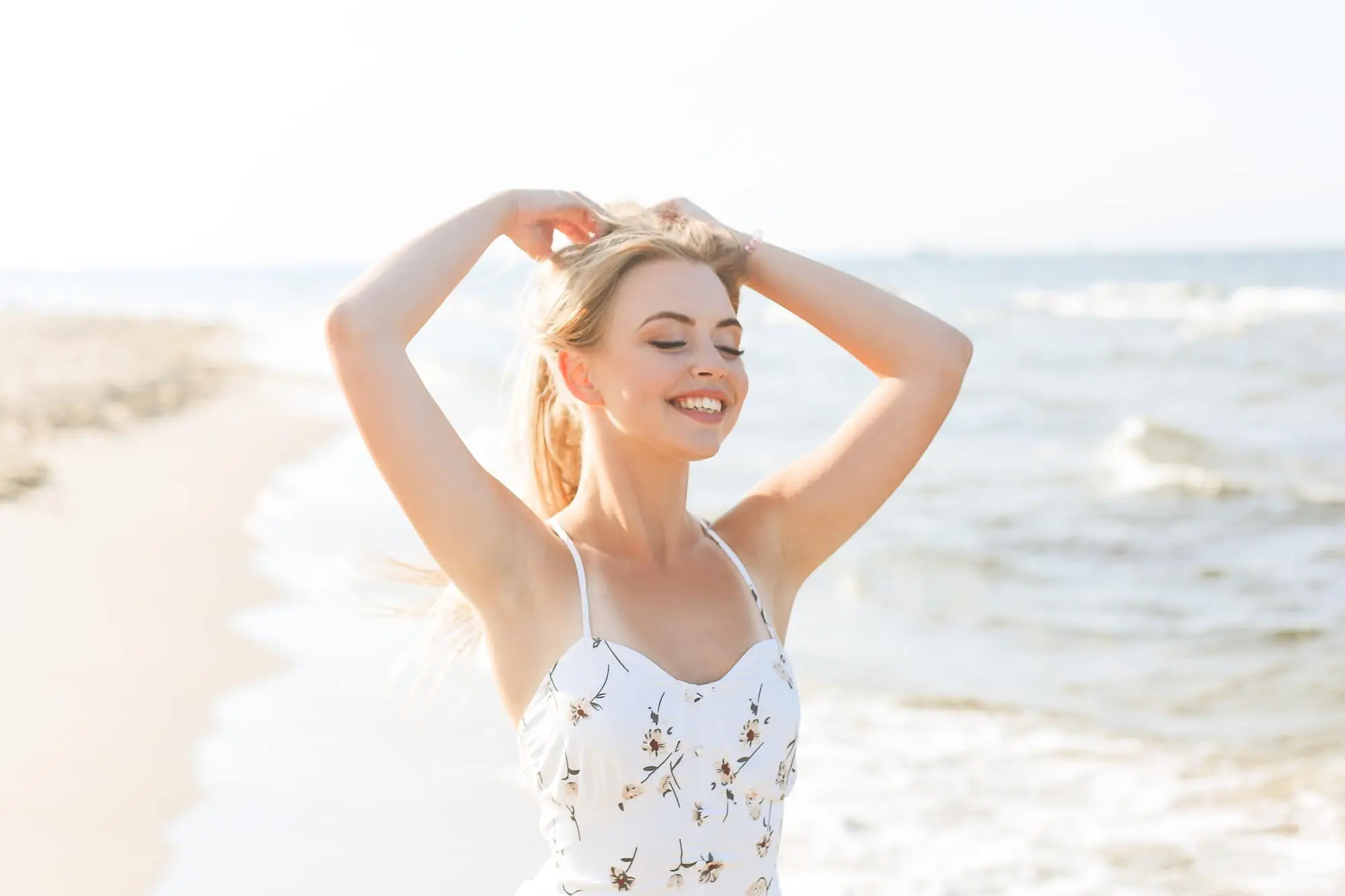 On a sunny beach in Cape Coral, FL, a woman glows with radiant skin by Refresh MedSpa, smiling in a white floral dress.