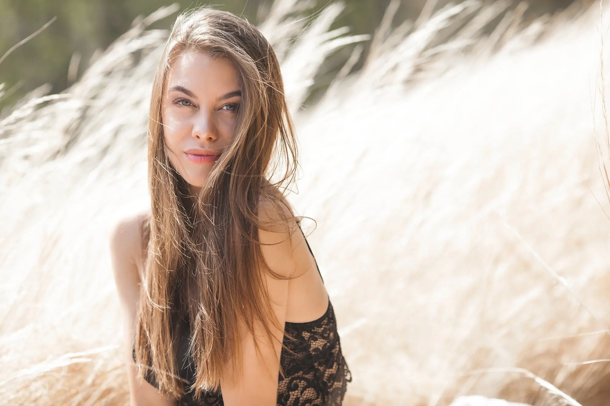 In Cape Coral, FL, a woman with long brown hair sits in tall dry grass, smiling at the camera for Refresh MedSpa.