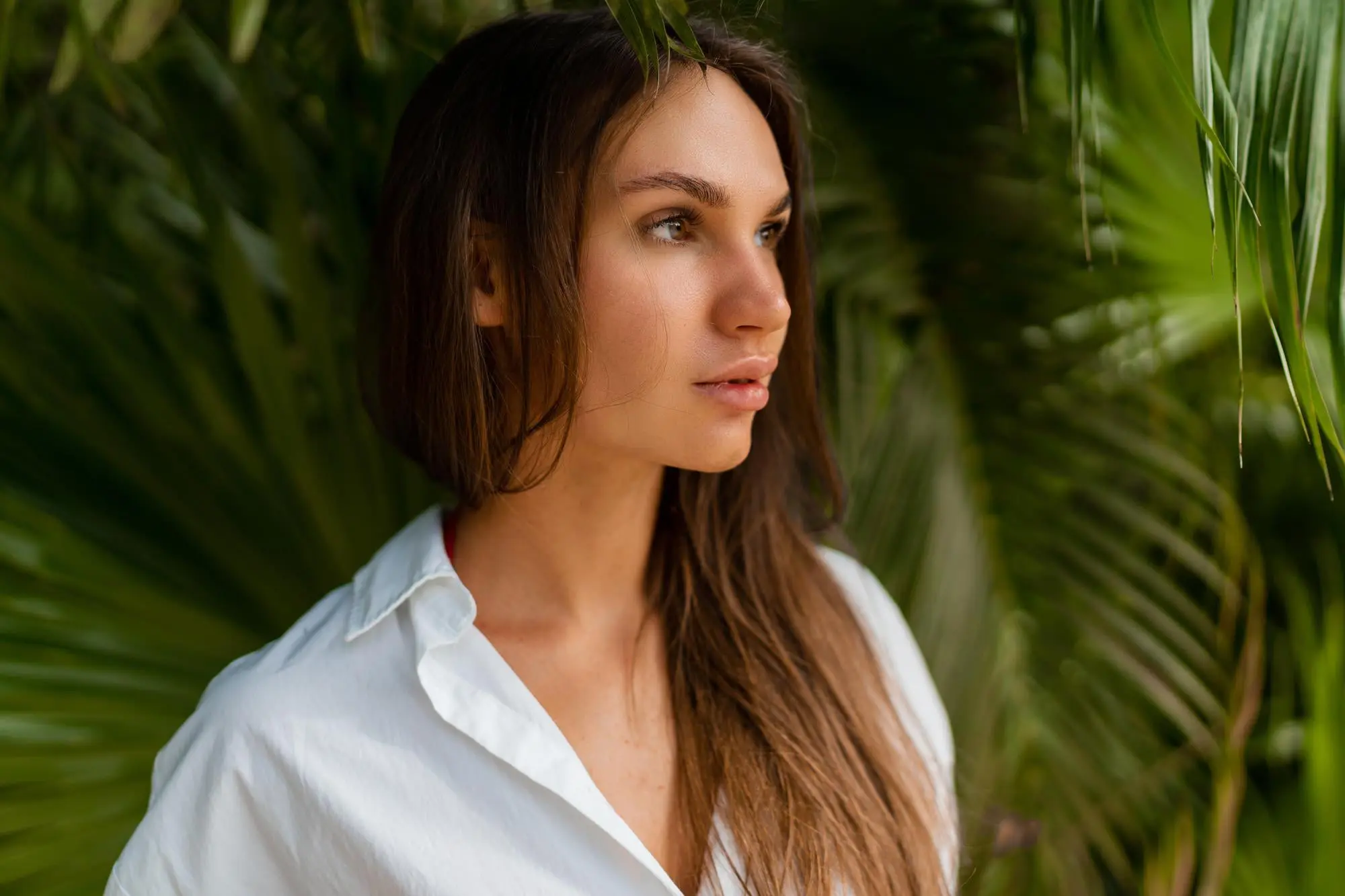 A woman with long brown hair, smooth skin, and a white shirt stands by lush palm leaves at Refresh MedSpa.