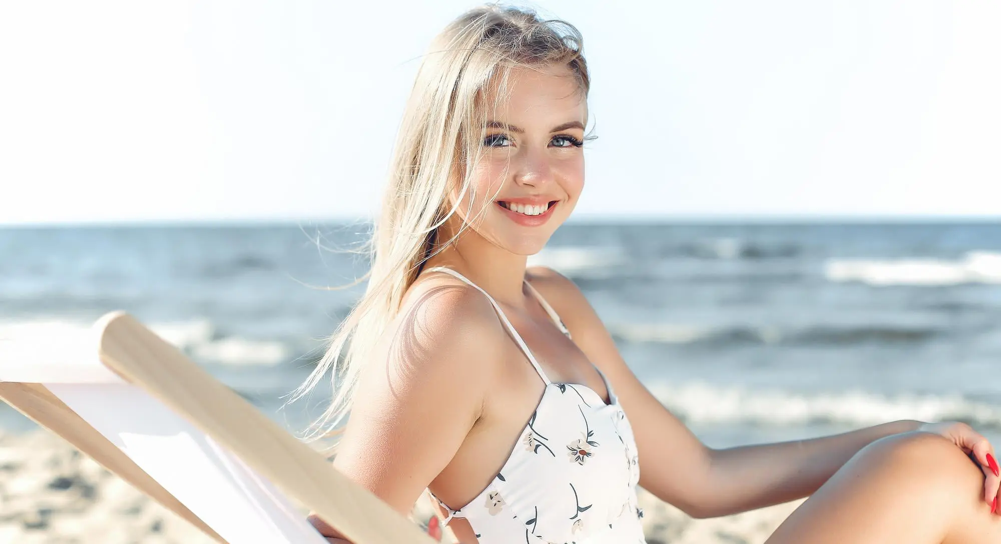 A young woman with smooth skin smiles on a beach lounge chair, ocean behind her, representing Refresh MedSpa in Cape Coral, FL.