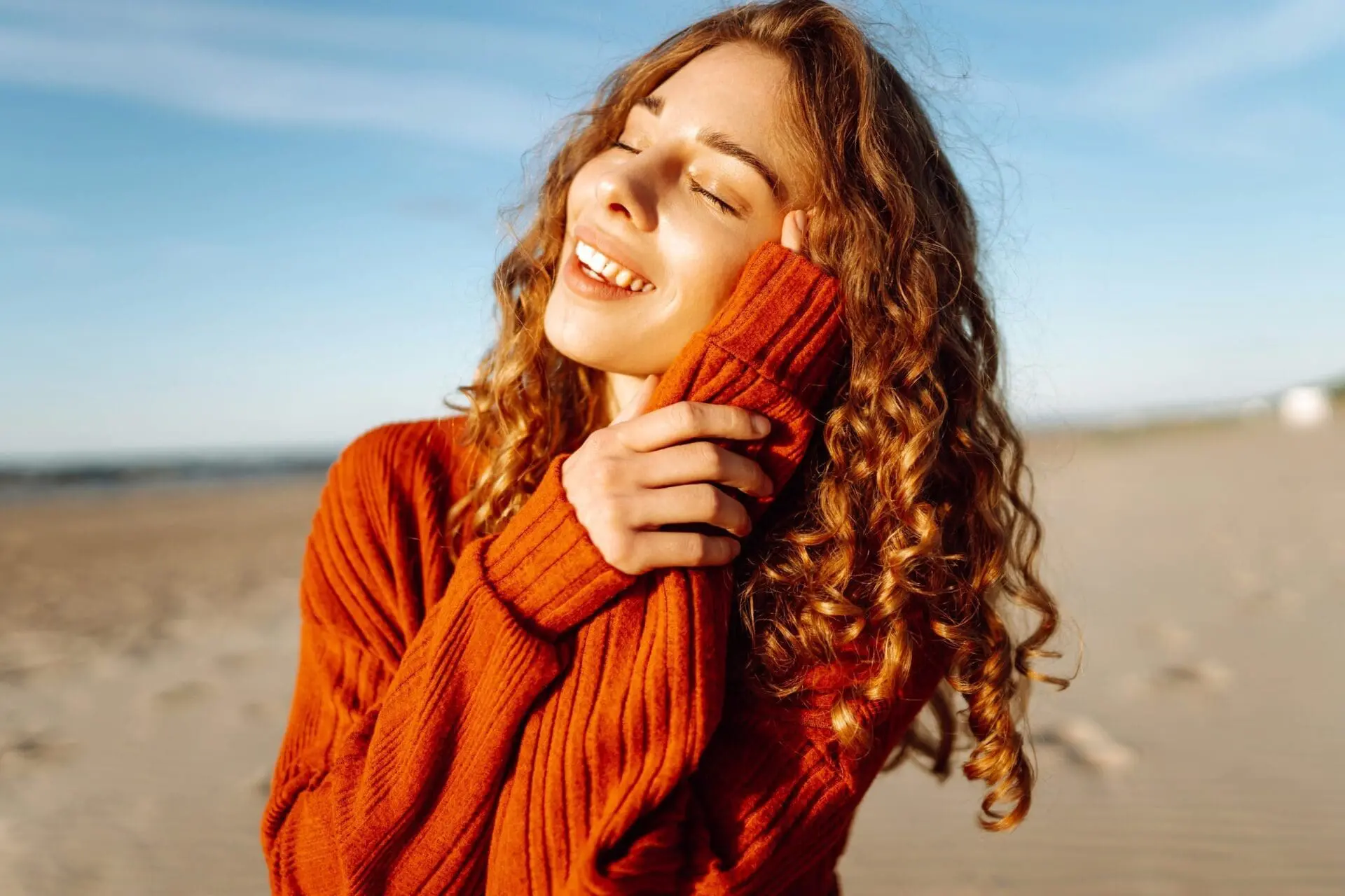 Smiling woman with curly hair in orange sweater on sandy beach, confident after rosacea treatment at Refresh MedSpa in Cape Coral, FL.