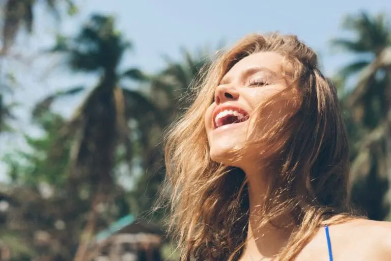 A woman with long hair smiles with her eyes closed, enjoying the sunlight outdoors near palm trees, radiating the glow of a recent med spa in Cape Coral, FL.