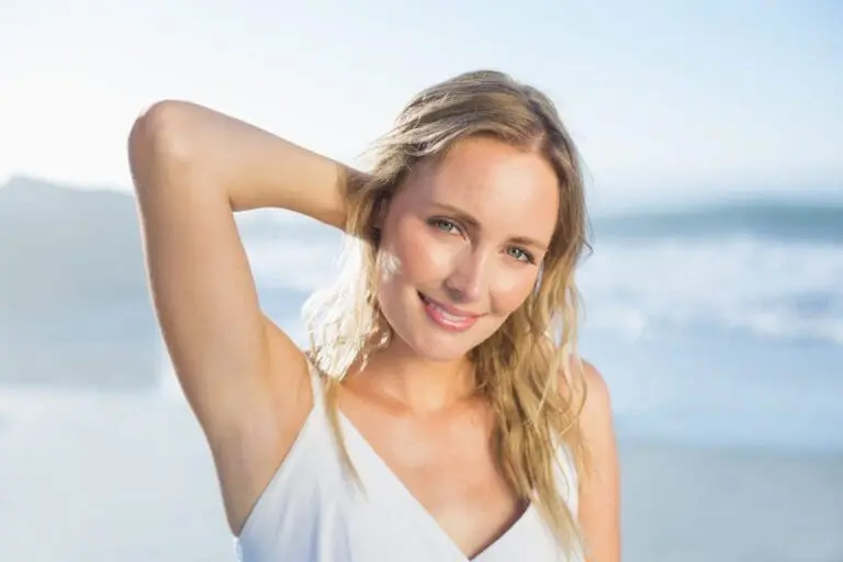 A woman with blonde hair stands on a beach near med spa in Cape Coral, smiling at the camera with one hand behind her head. She is wearing a white sleeveless top and the ocean is visible in the background.