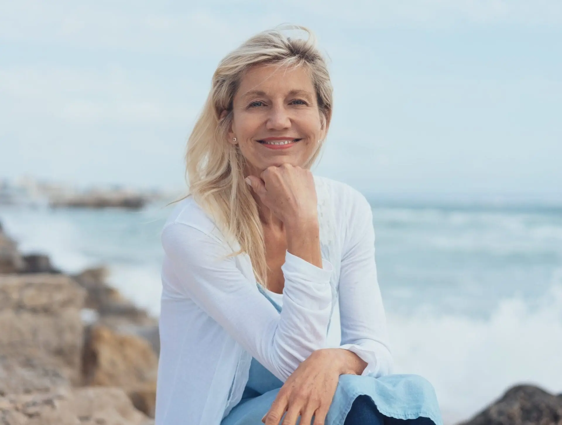 A woman with long blonde hair sits on rocks by the sea, smiling and resting her chin on her hand, with waves and a cloudy sky in the background.