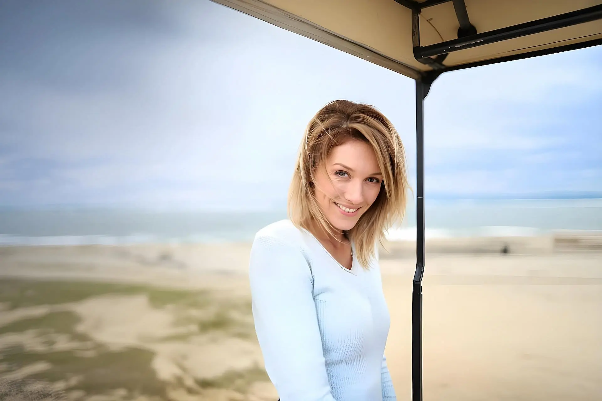 A woman with shoulder-length blonde hair stands under a canopy near a beach in Cape Coral, smiling at the camera with the ocean and sand behind her—showcasing the radiant results of botox Cape Coral FL.