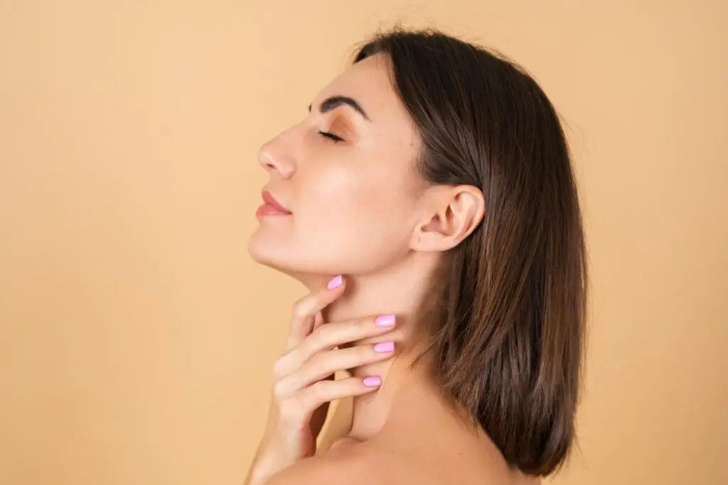 At Refresh MedSpa in Cape Coral, FL, a woman with short brown hair stands in profile against a beige background, eyes closed and gently touching her neck, her features softly enhanced by cheek fillers.