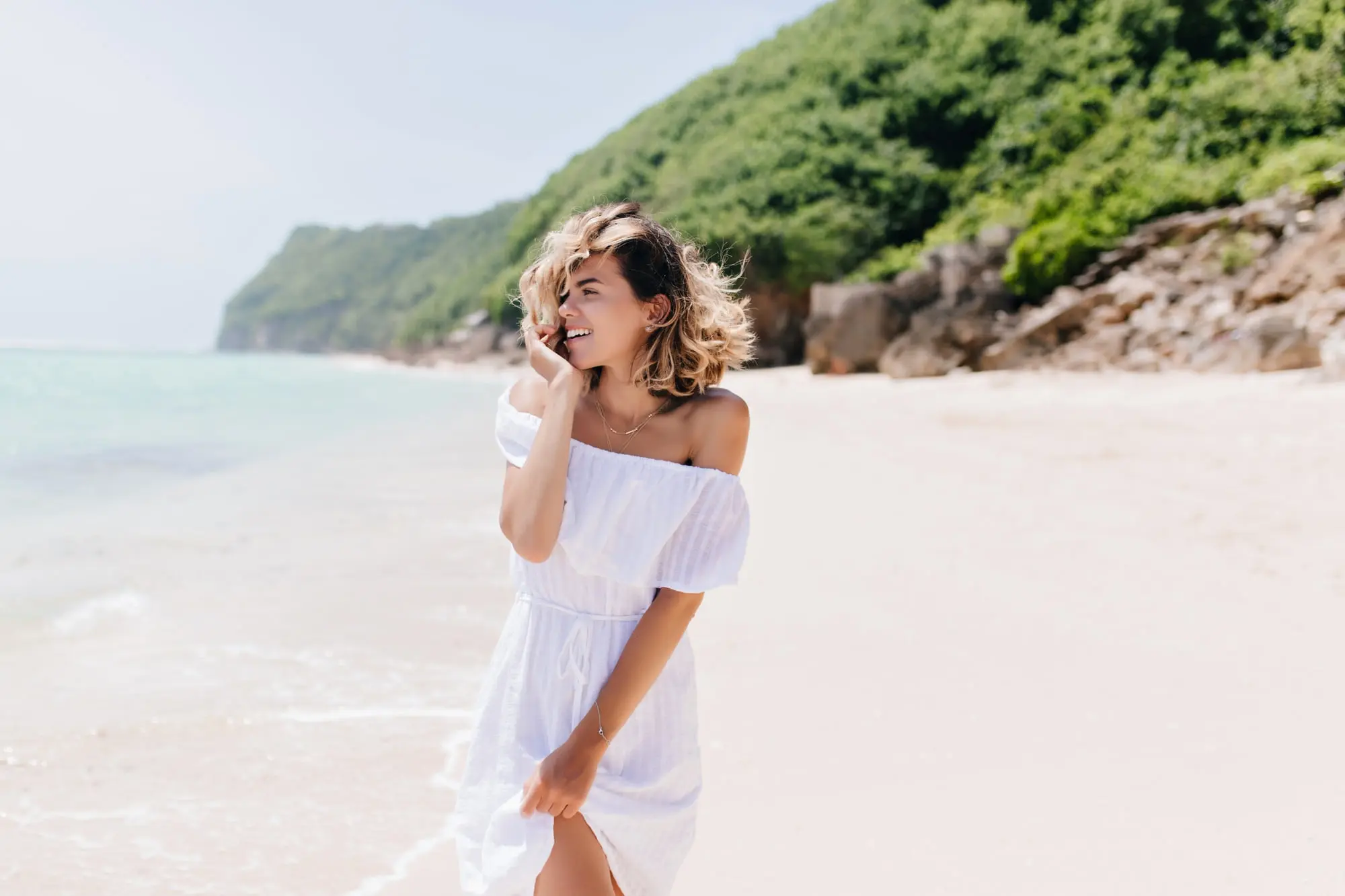 On a sandy beach, a woman in white smiles on the phone, showing Botox benefits from Refresh MedSpa.