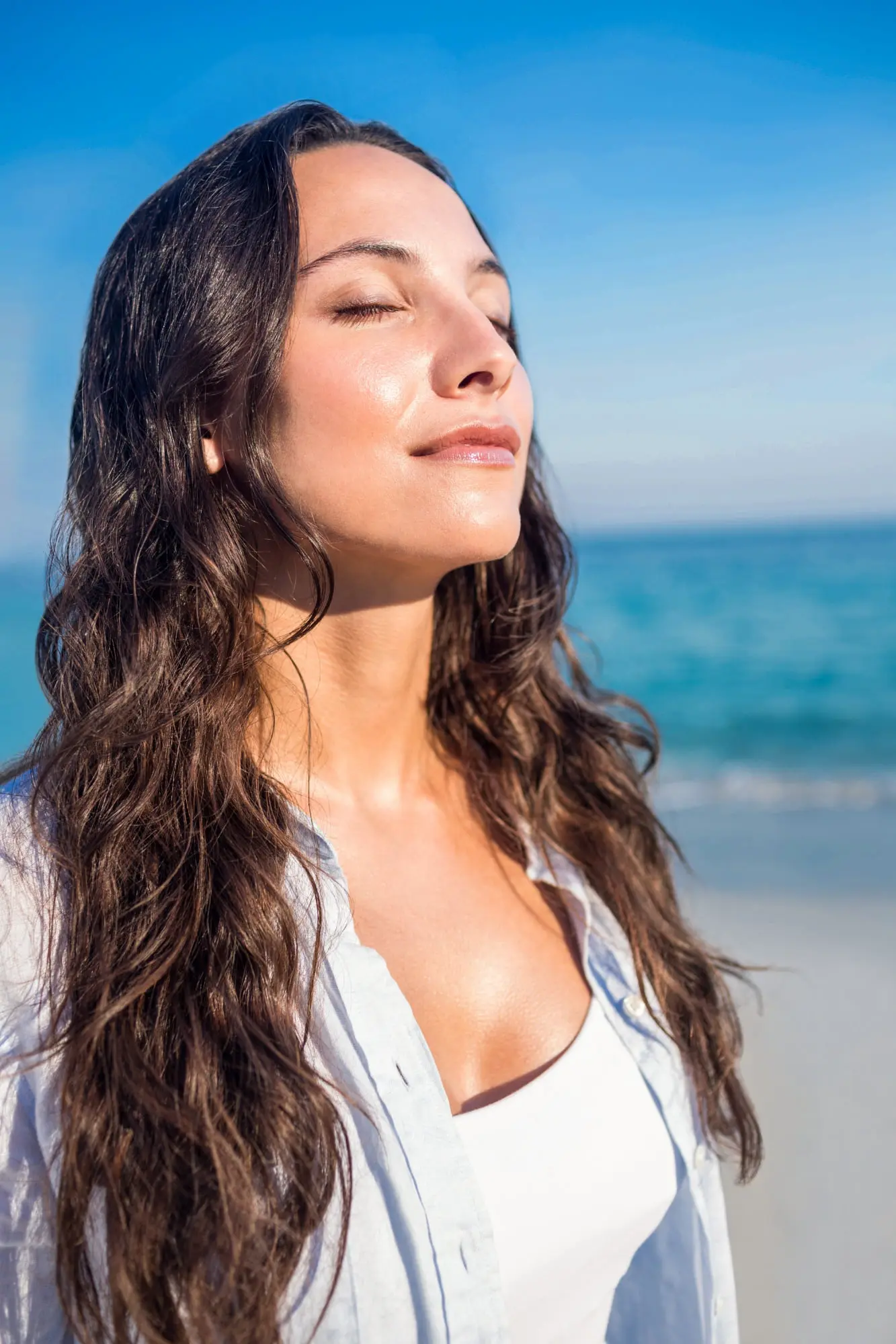 A woman with long brown hair enjoys the sun after Juvederm at Refresh MedSpa; sky and ocean behind.