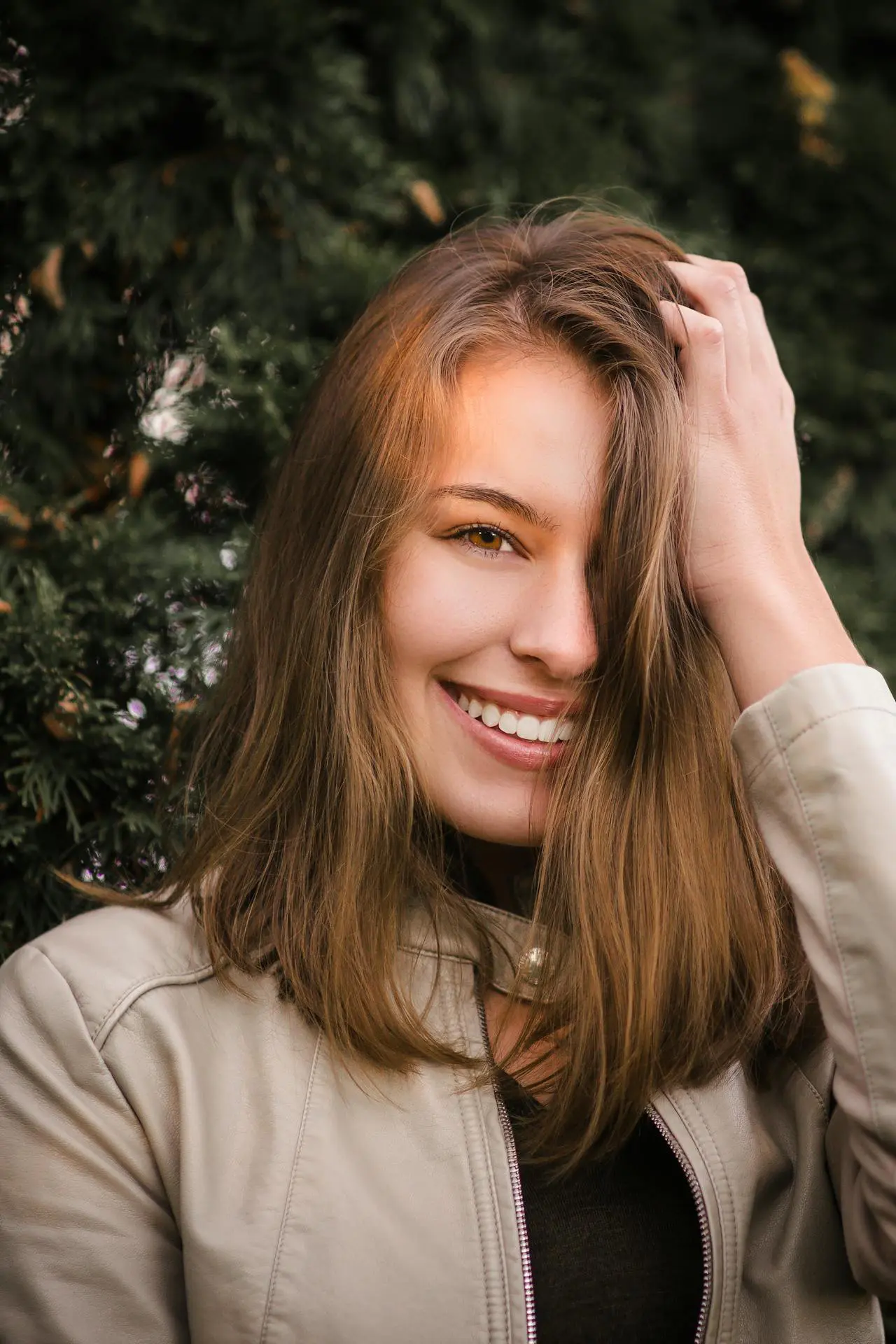 Smiling in front of green foliage, a young woman in a light jacket shows smooth skin from natural Dysport at Refresh MedSpa.