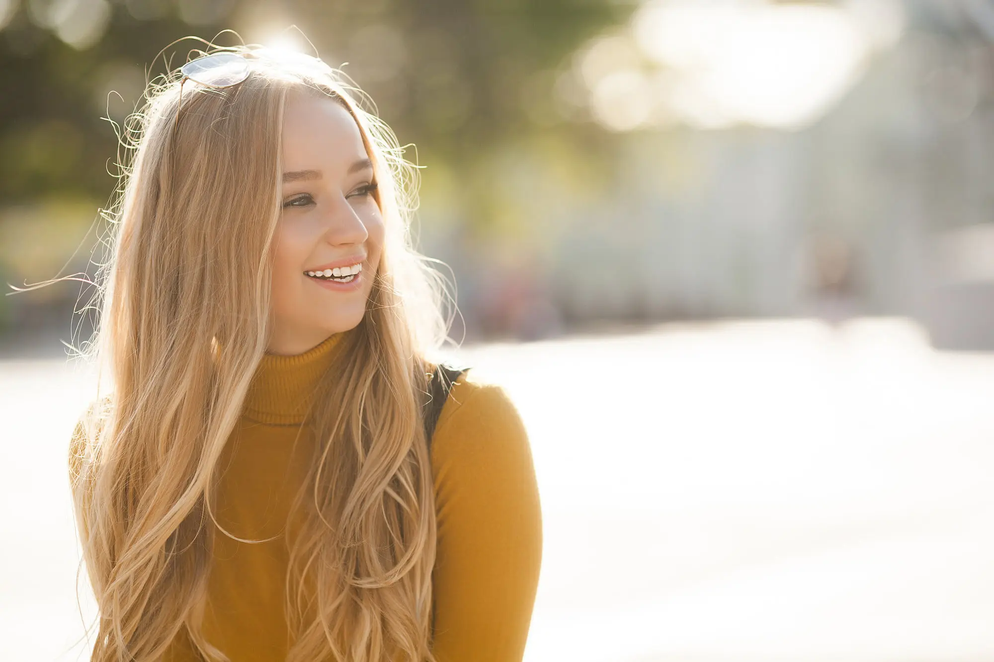 Smiling outdoors in bright light, a young blonde woman in a mustard sweater glows after Forever Clear BBL at Refresh MedSpa.