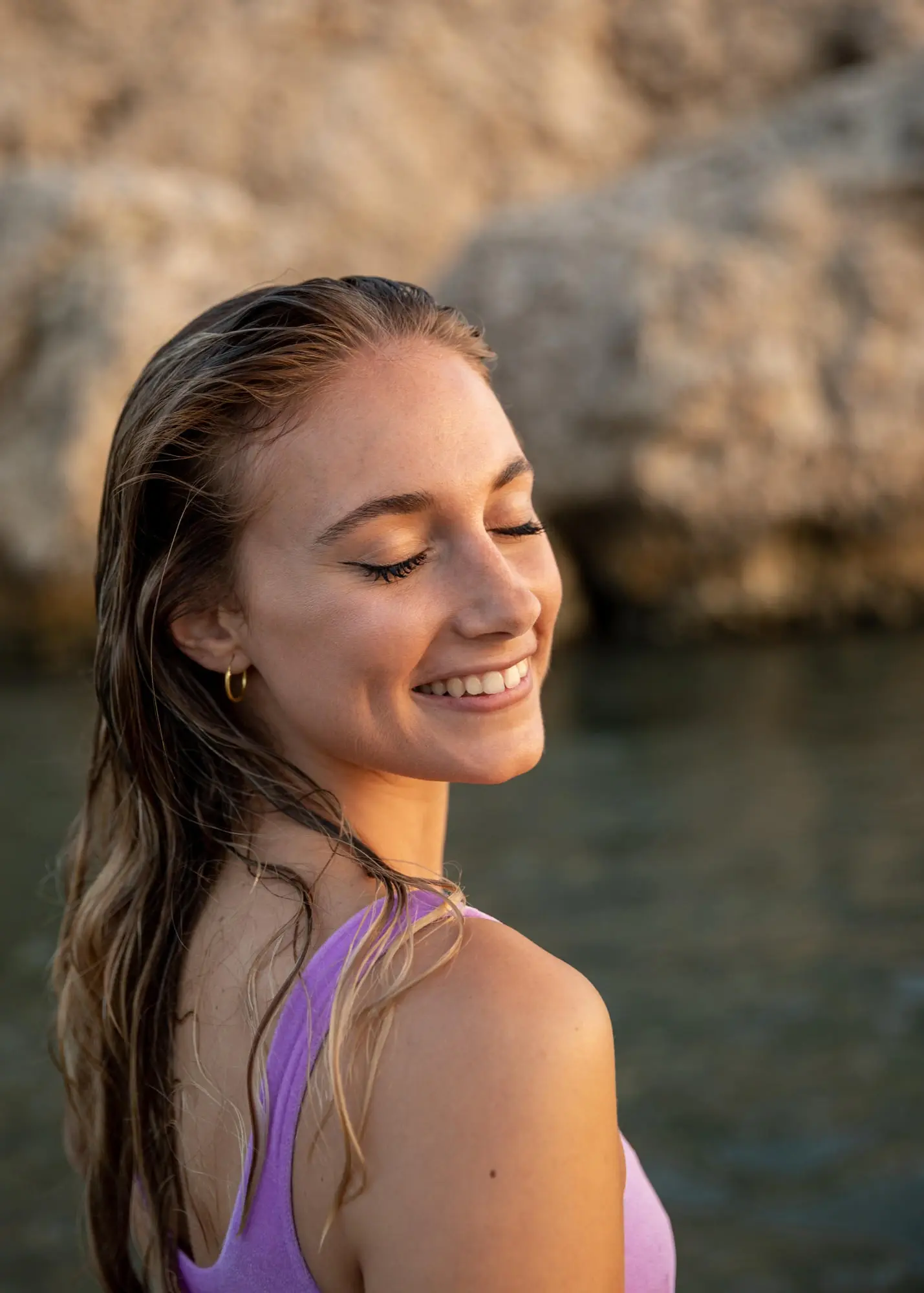Smiling with closed eyes, a woman in a purple top stands by water and cliffs, glowing after Radiesse at Refresh MedSpa.