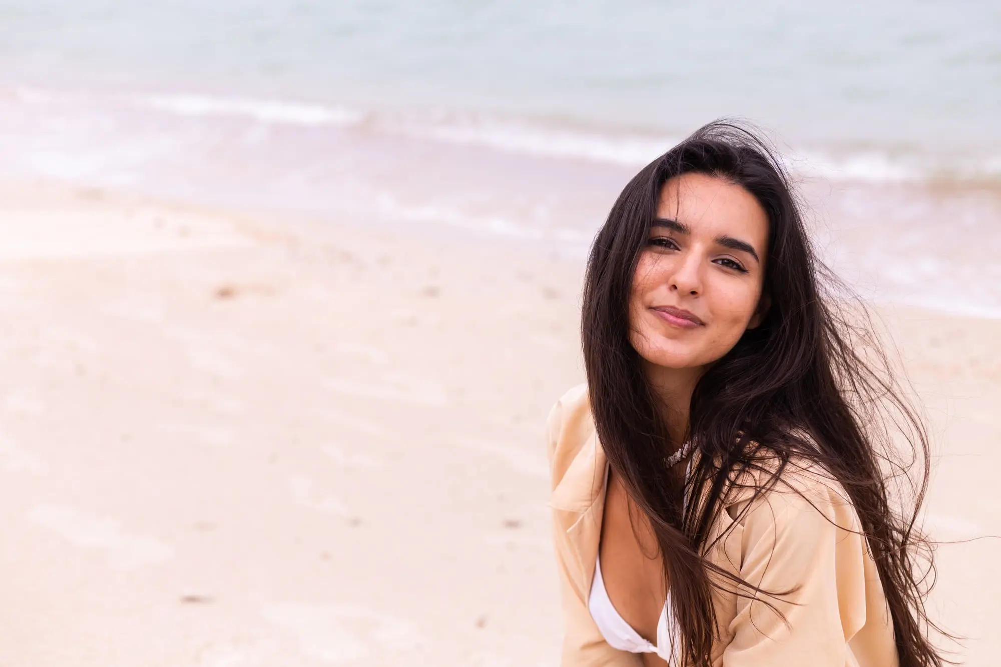 A woman with long dark hair smiles on a sandy beach, her fresh look showing Sculptra results from Refresh MedSpa.