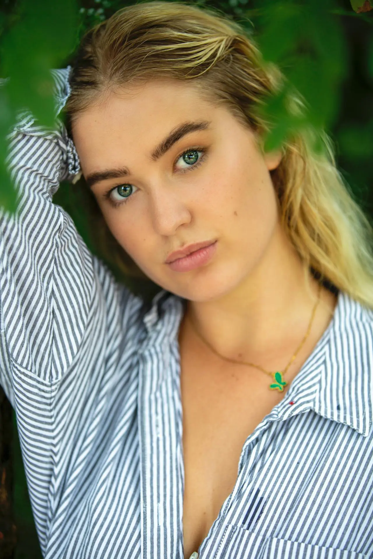 A refreshed person with long blond hair, blue eyes, and a striped shirt poses by green leaves after Xeomin at Refresh MedSpa.