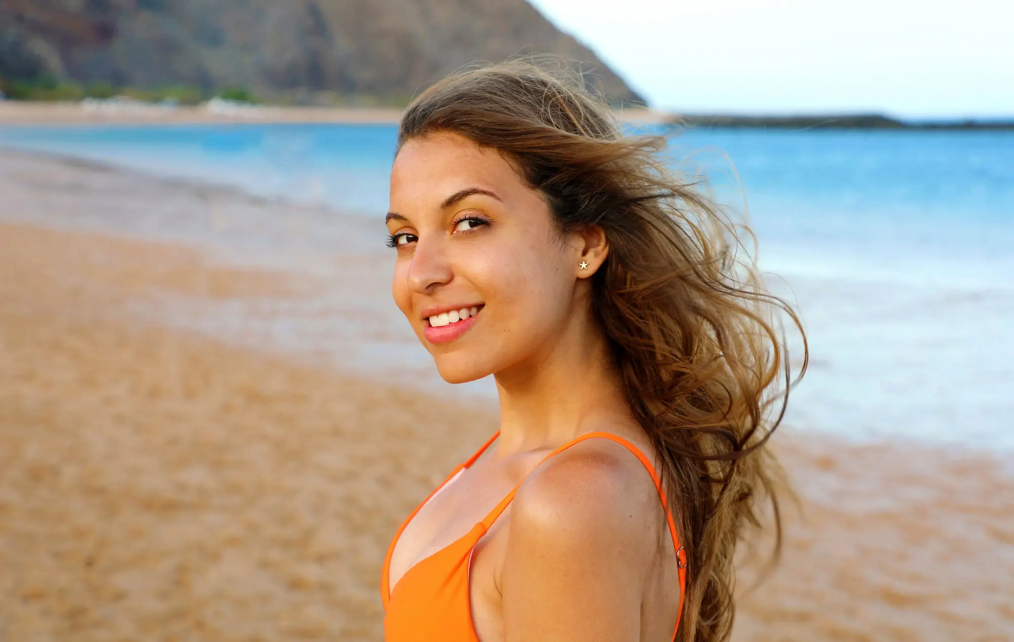 A woman in an orange swimsuit smiles on a beach, glowing after PRP Skin Rejuvenation at Refresh MedSpa.