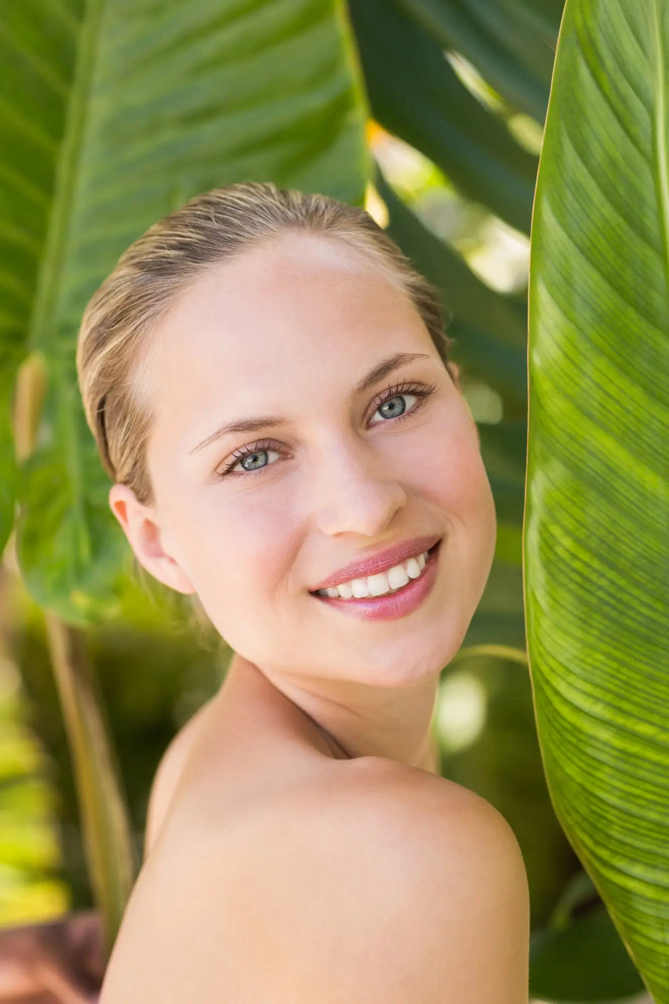 A blonde woman smiles outdoors by lush tropical leaves, her skin glowing after a BBL at Refresh MedSpa.