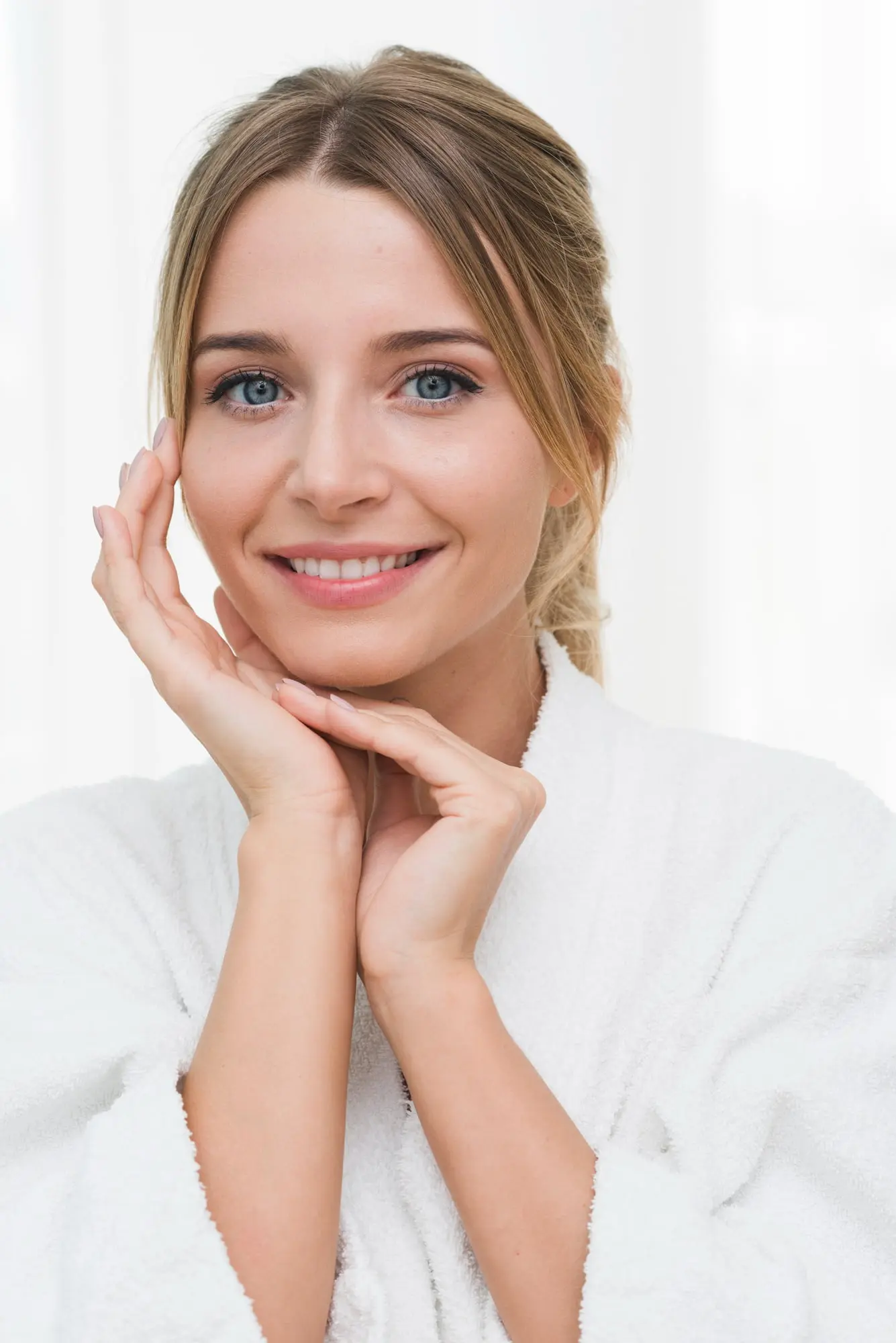 A blonde woman in a white bathrobe smiles, touching her face after a BBL photofacial at Refresh MedSpa.
