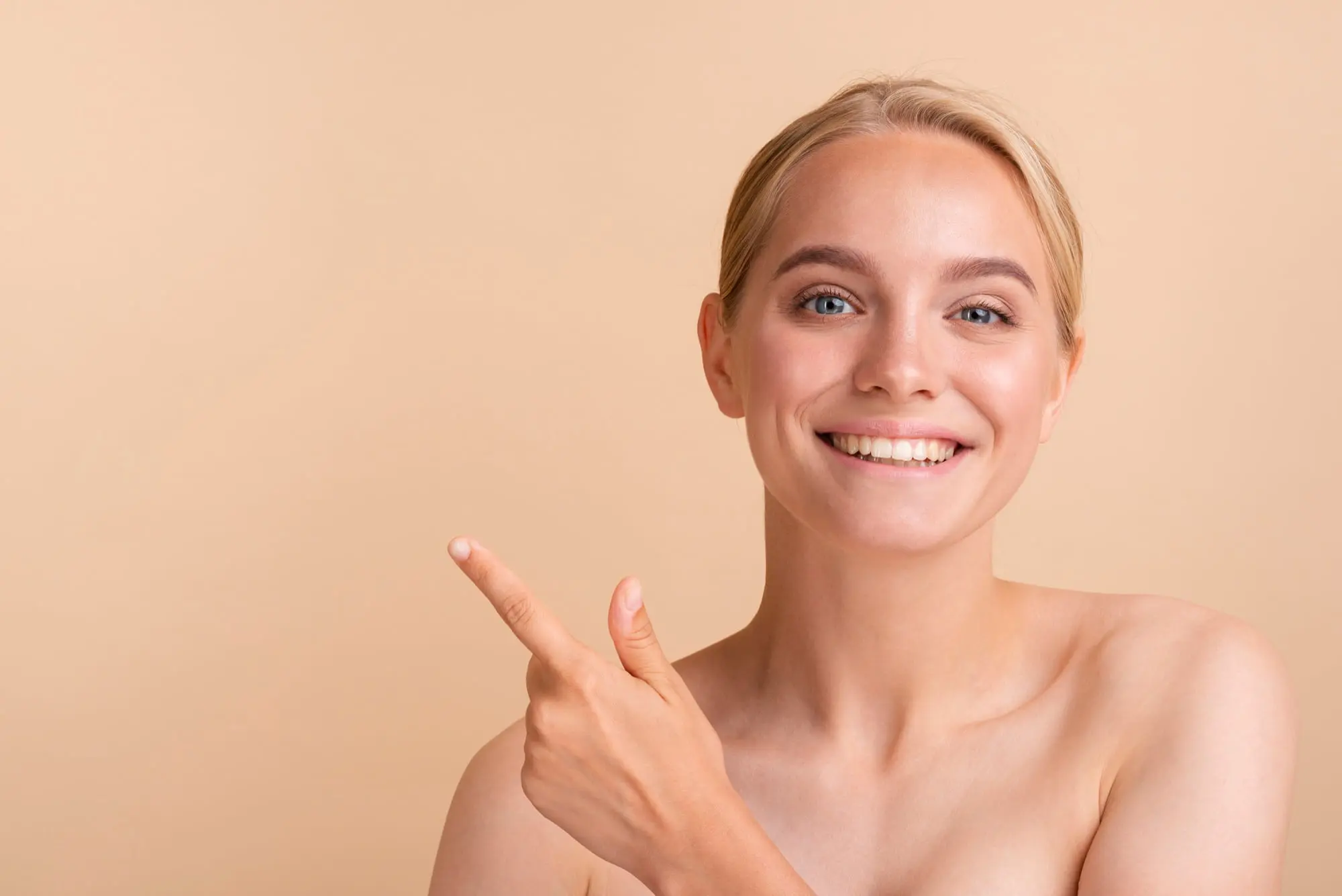 Confident, bare-shouldered blonde woman points left, smiling after a BBL photofacial at Refresh MedSpa.