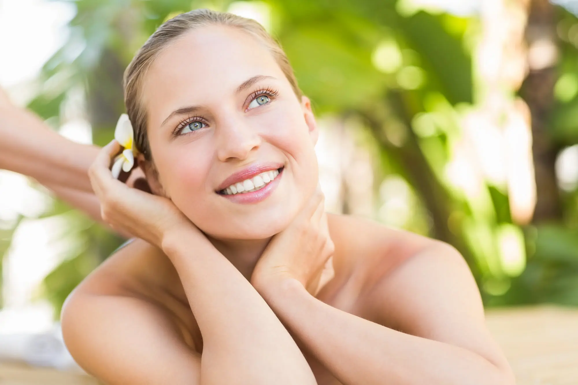 A woman at Refresh MedSpa, smiles as a white flower is placed behind her ear, her skin glowing amid green foliage.