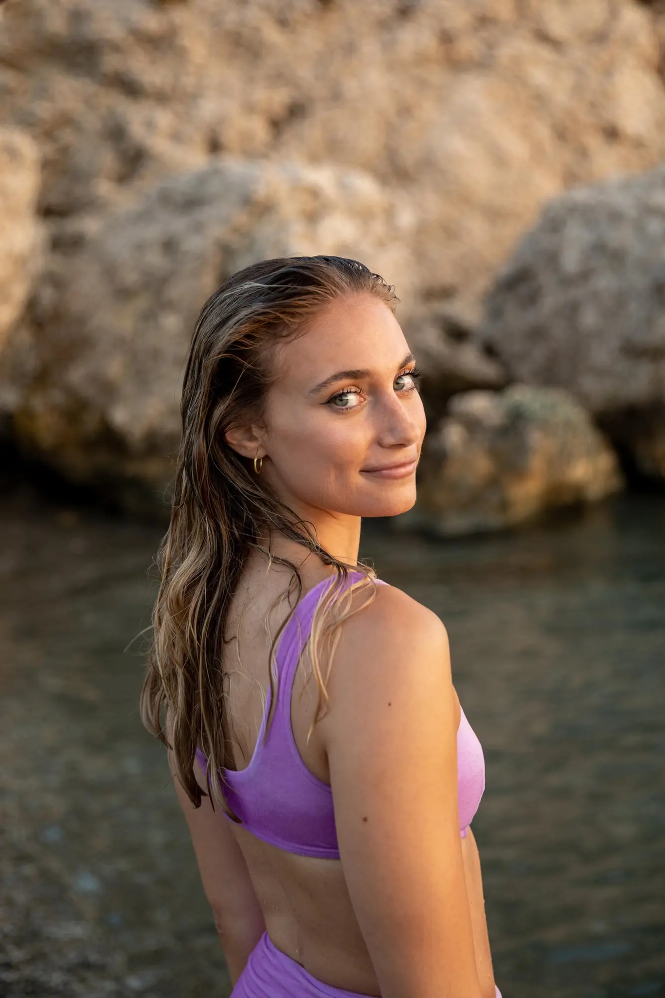 By rocky water, a woman in a purple bikini shows glowing HydraFacial skin from Refresh MedSpa.