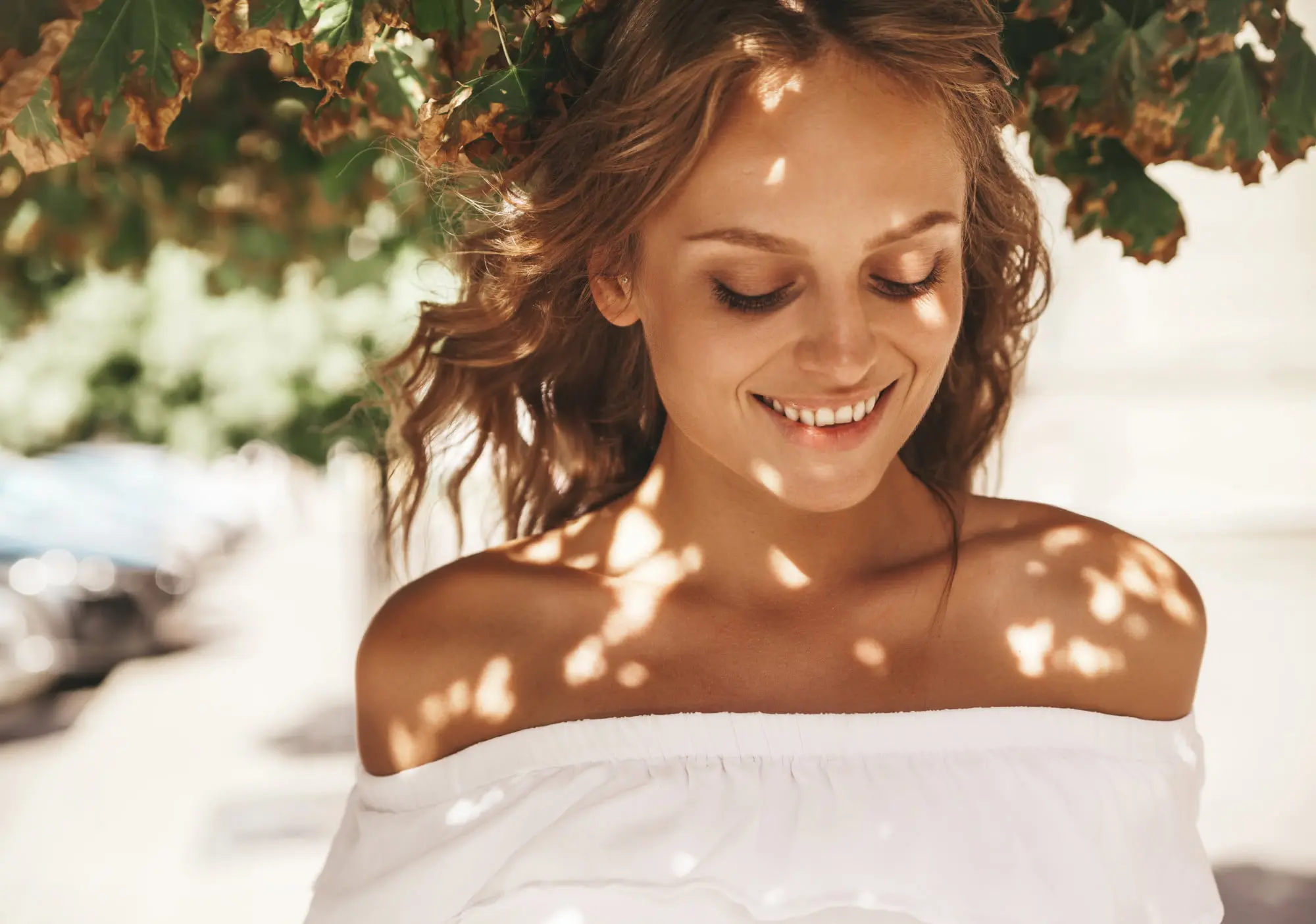 Smiling woman in an off-shoulder white top stands under leafy branches, glowing after a HydraFacial at Refresh MedSpa.