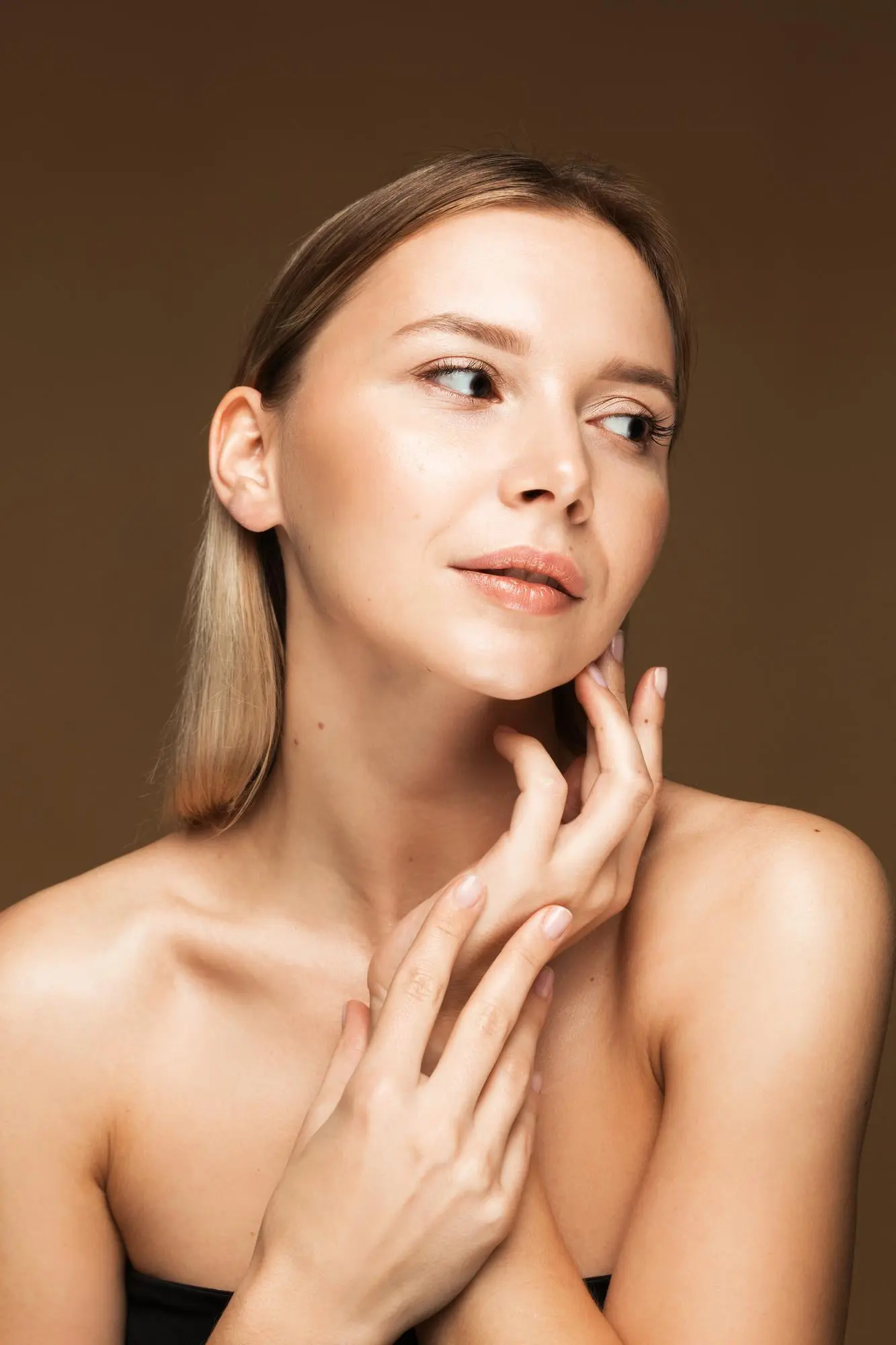 A woman with glowing skin from a HydraFacial at Refresh MedSpa, poses against a brown background, touching her face.