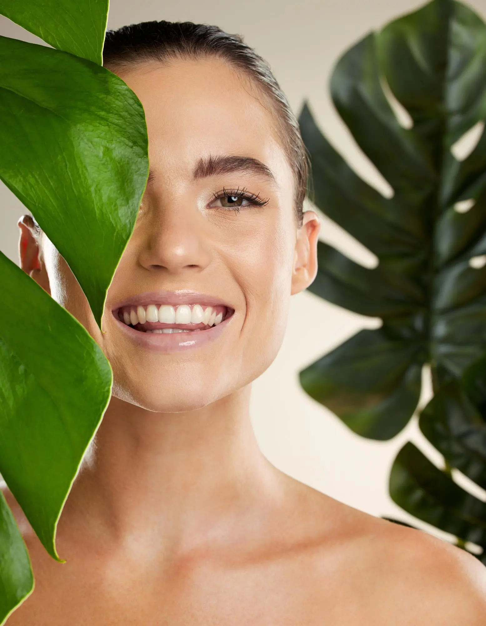 At Refresh MedSpa, a woman smiles with moxi, her face partly hidden by a large green leaf among lush plants.