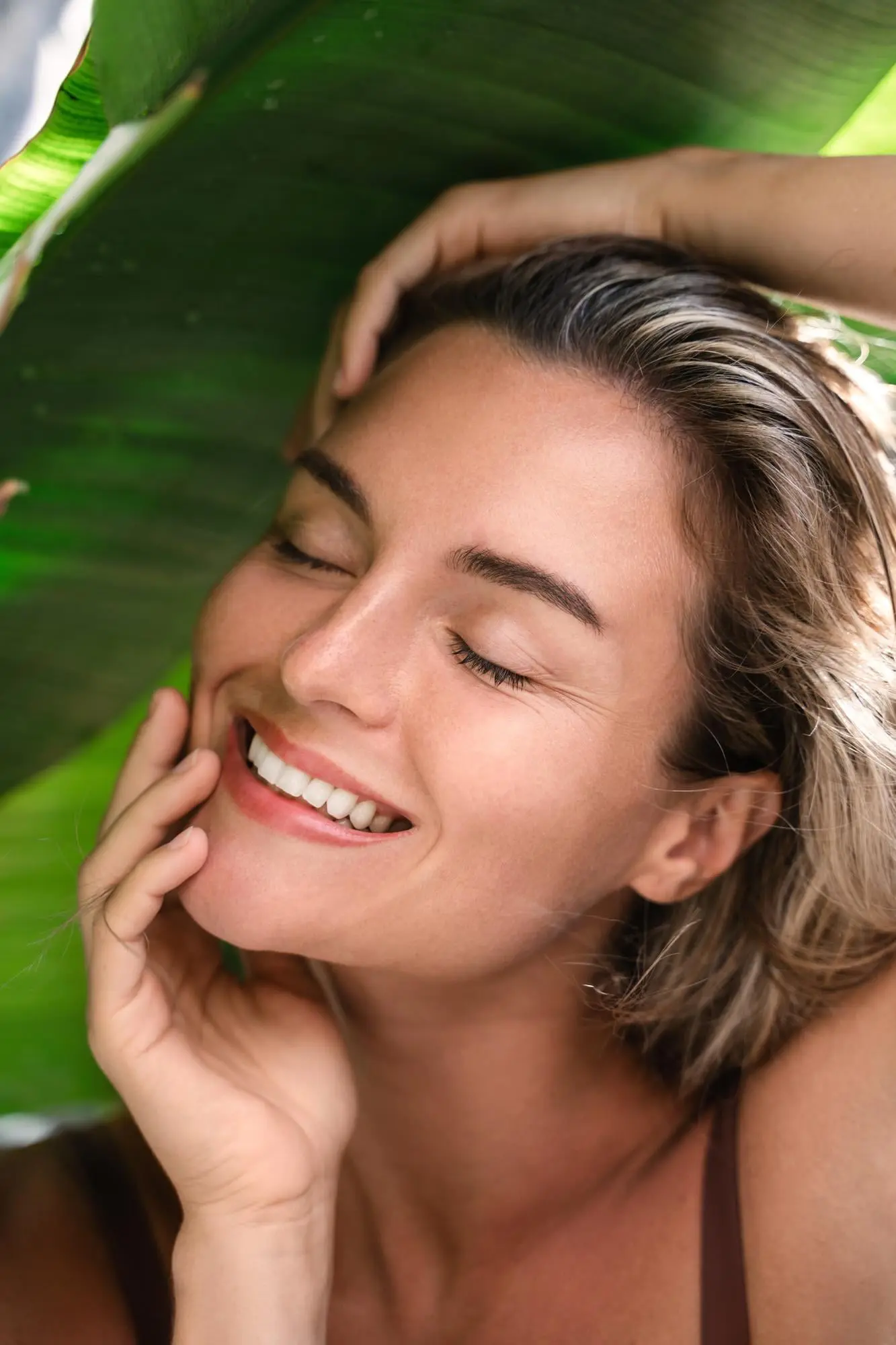 Under a large green leaf, a serene person smiles softly at Refresh MedSpa, gently touching their face.