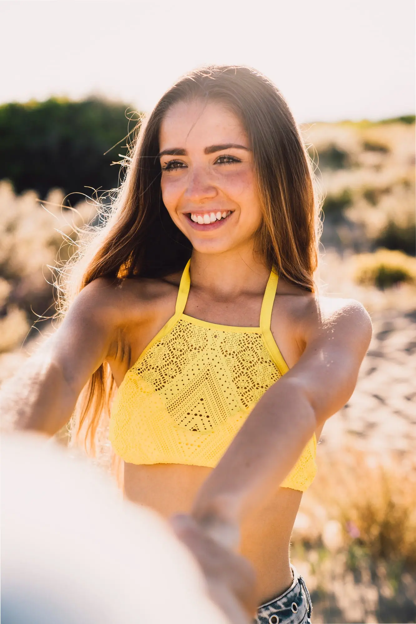 Smiling outdoors on a sunny day, a young woman in a yellow halter top holds someone's hand at Refresh MedSpa.