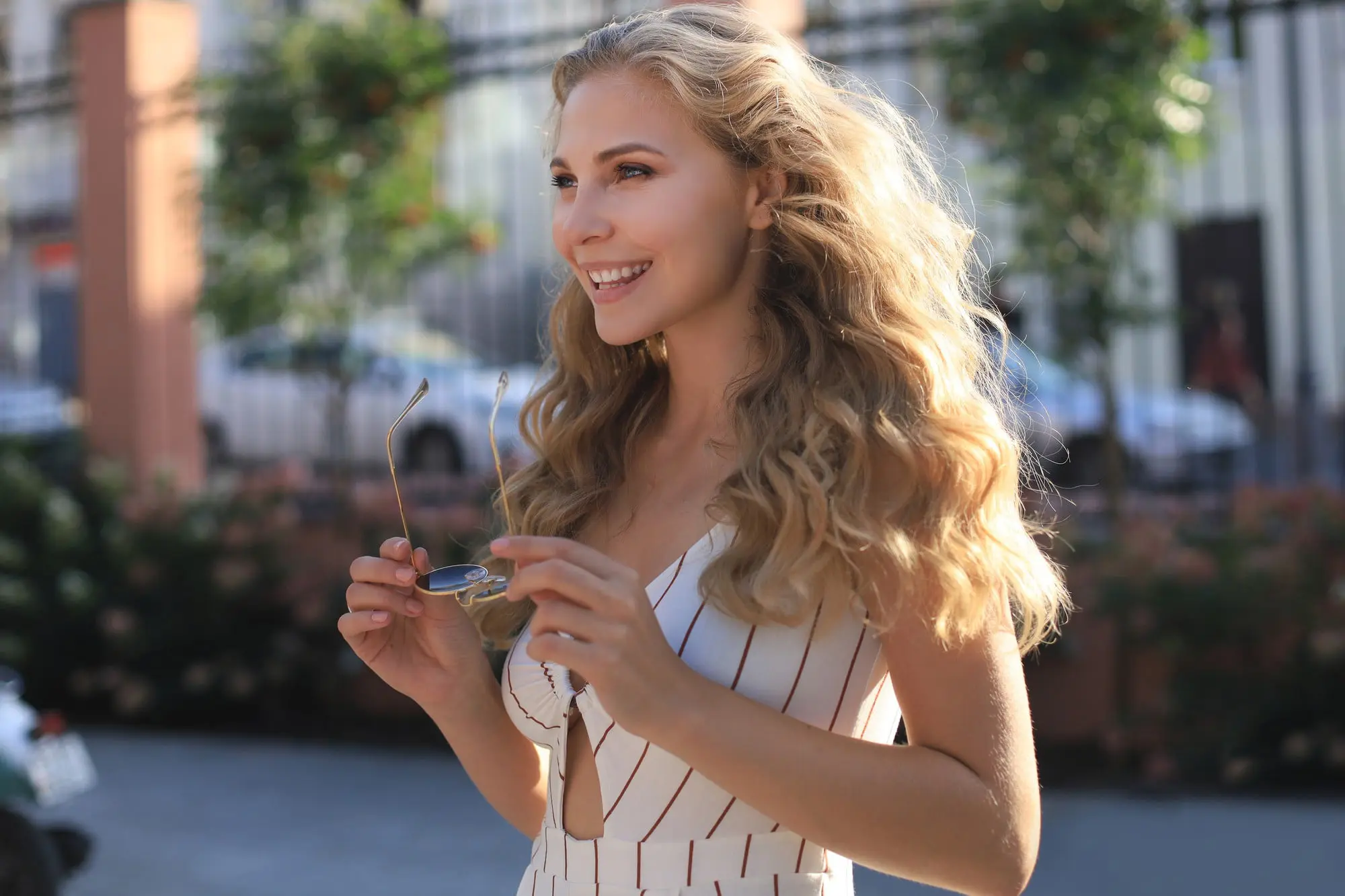 Smiling woman with long blonde hair and glowing skin from Moxi + BBL at Refresh MedSpa, holds sunglasses outside.