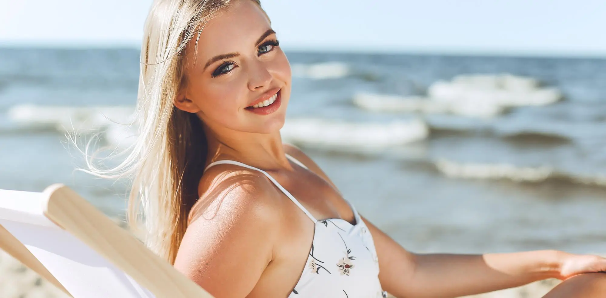 A blonde woman in a white swimsuit sits by the sea with glowing skin, showcasing Refresh MedSpa results.