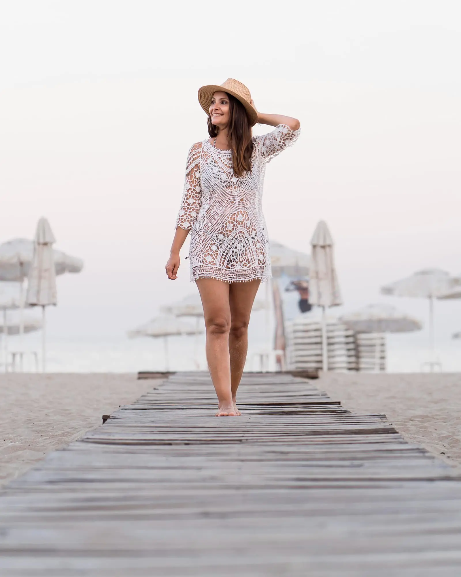 At Refresh MedSpa in Cape Coral, FL, a Beauty Bank member in a white lace cover-up and straw hat walks barefoot on a beach path.