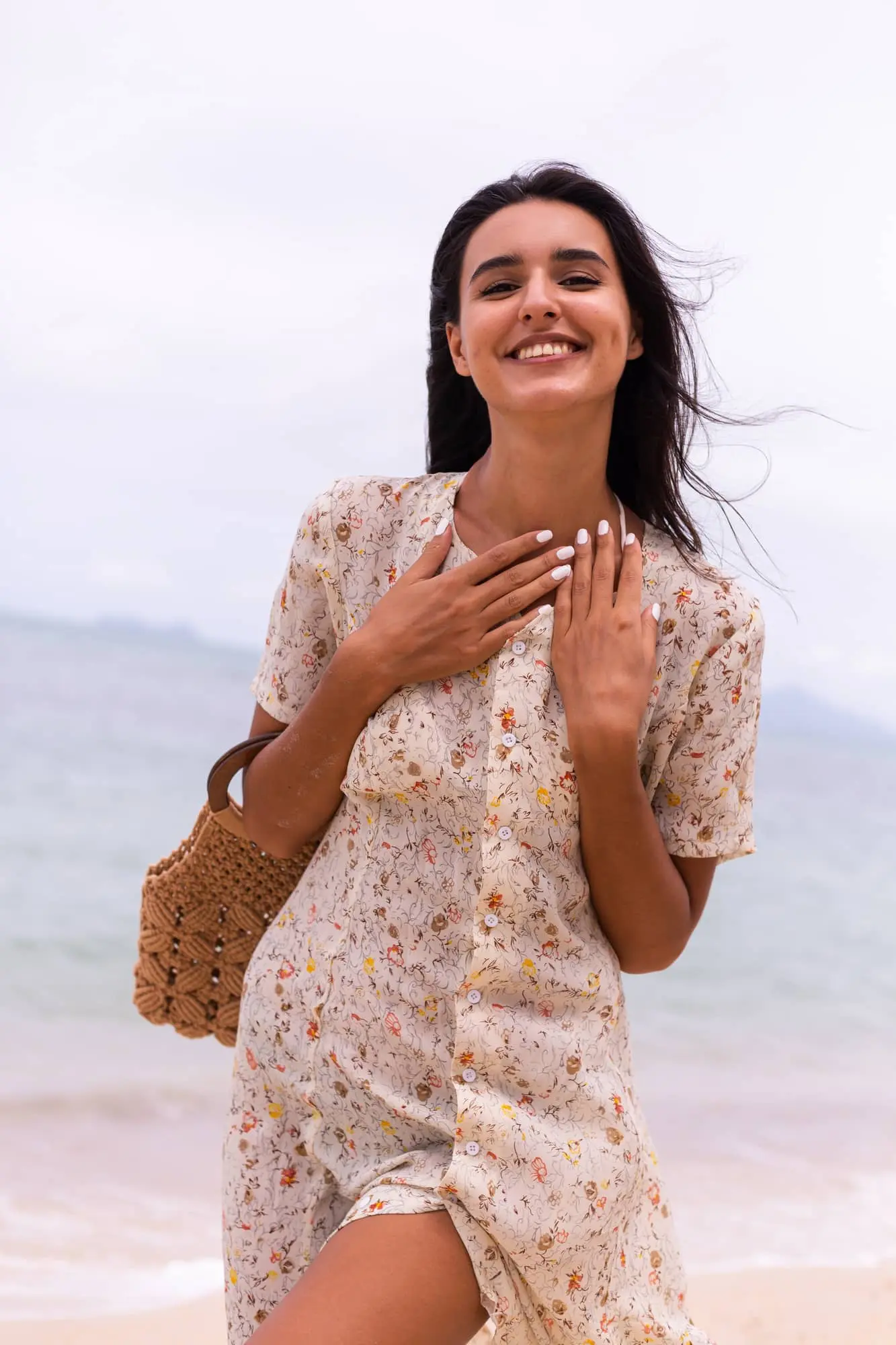 On a Cape Coral, FL beach near Refresh MedSpa, a smiling woman in a floral dress holds a woven bag, with sea and sky behind her.