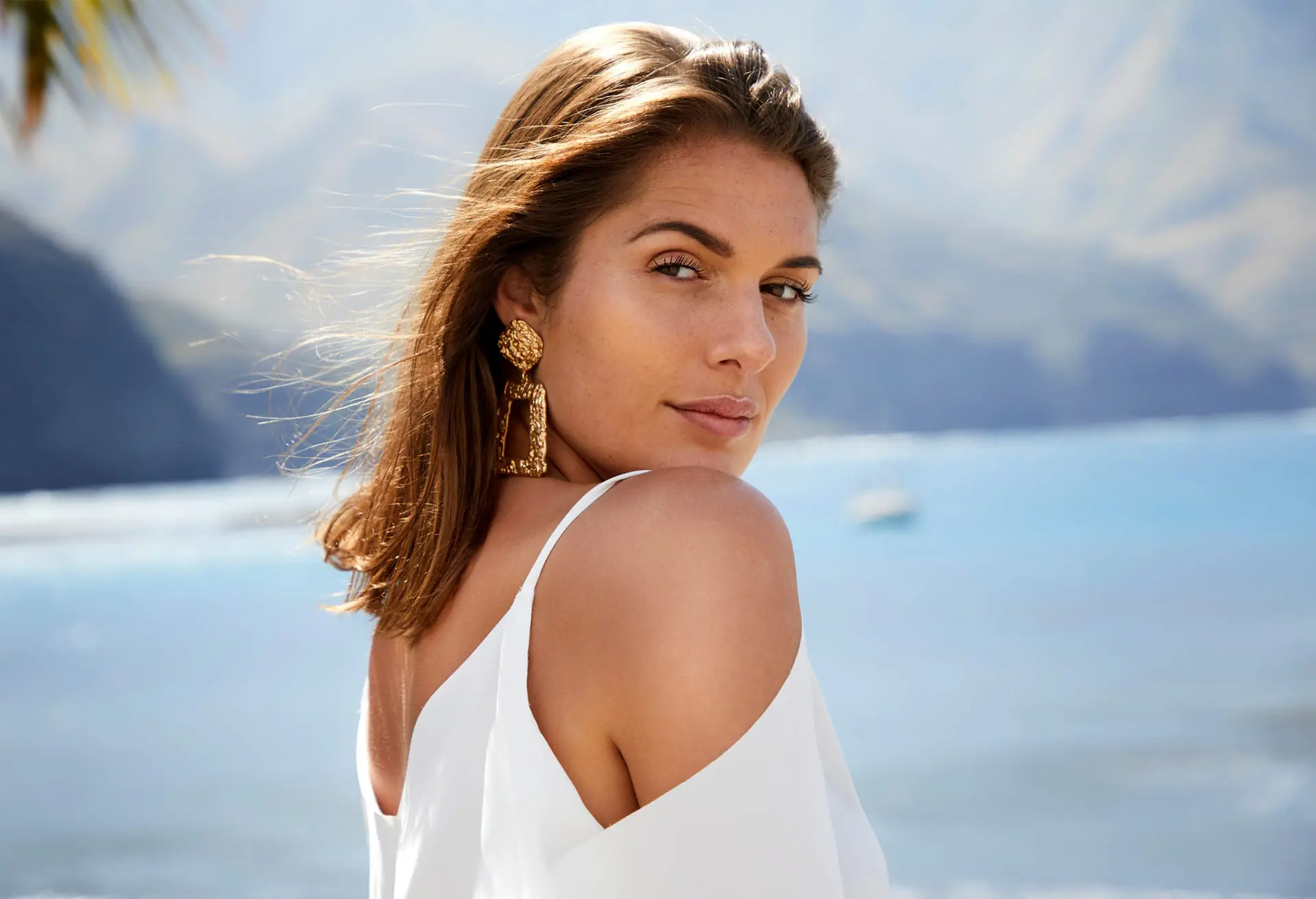 Woman with shoulder-length brown hair wearing a white top and gold earrings stands outdoors near a body of water with mountains in the background, reminiscent of the tranquility found after enjoying lasers in Cape Coral, FL.