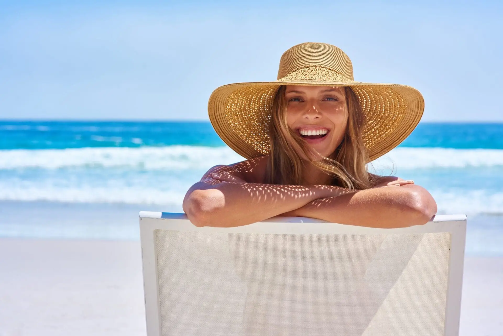 Smiling woman in wide-brimmed hat on a beach chair, her glowing skin shows results from Refresh MedSpa in Cape Coral, FL.