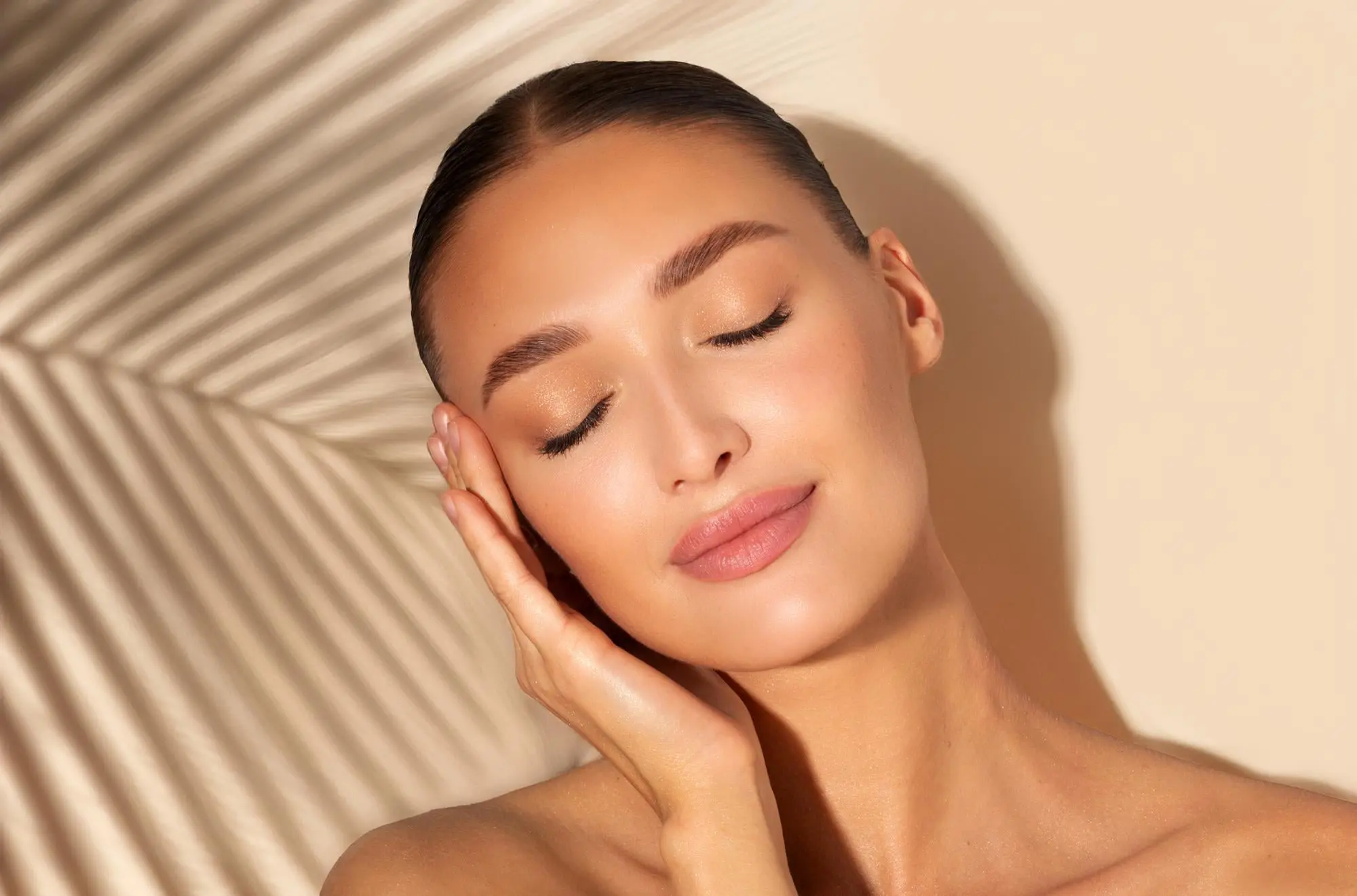 A woman with a gentle smile touches her face, basking in warm light at Refresh MedSpa in Cape Coral, FL after a HydraFacial.