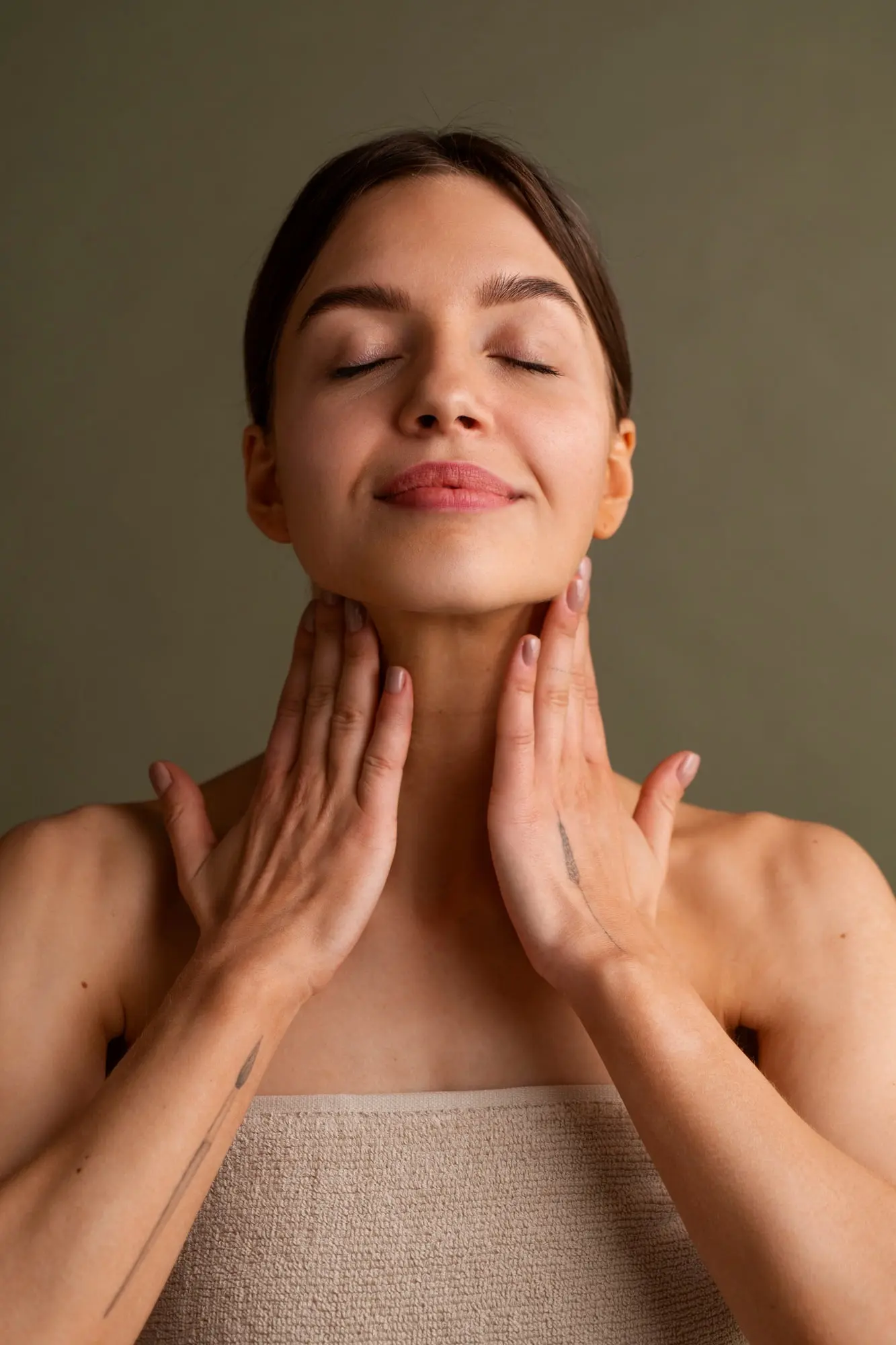 A woman with closed eyes gently touches her neck, wrapped in a beige towel—reflecting natural beauty at Refresh MedSpa in Cape Coral, FL.