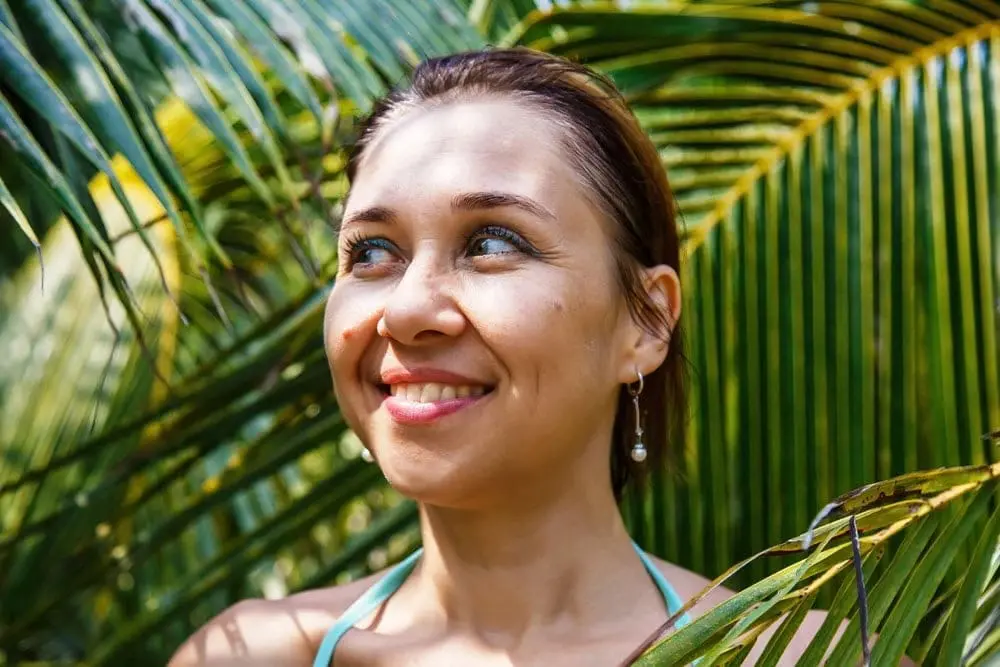 A woman smiles outdoors among palm leaves, sunlight on her face, reflecting the fresh confidence of Refresh MedSpa in Cape Coral, FL.
