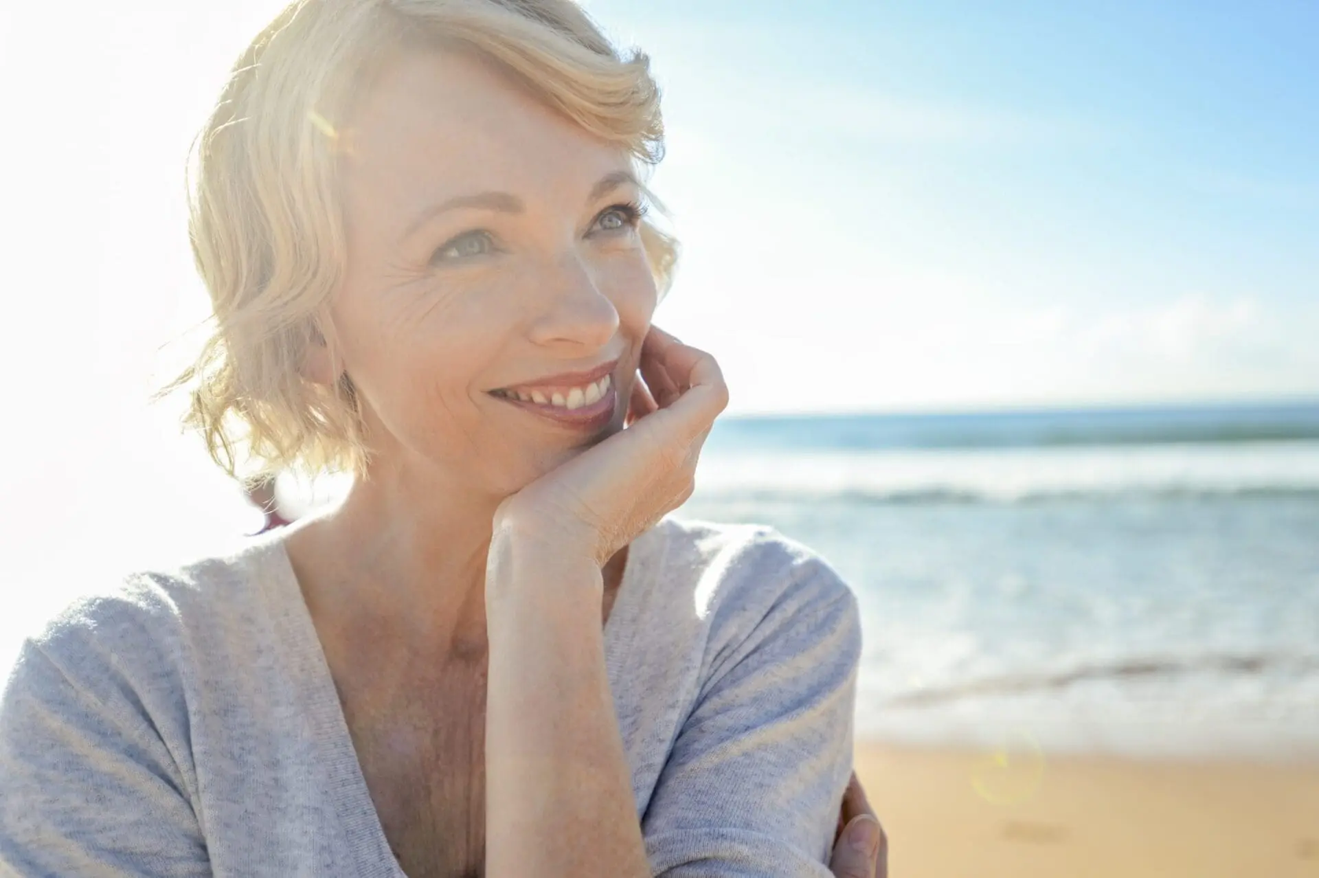 A confident, middle-aged woman with short blonde hair smiles on a sunny Cape Coral, FL beach after her Refresh MedSpa Radiesse treatment.