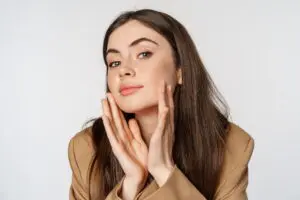 Young woman with long brown hair in a tan blazer touching her face, wondering how long Juvederm lasts at Refresh MedSpa in Cape Coral, FL.