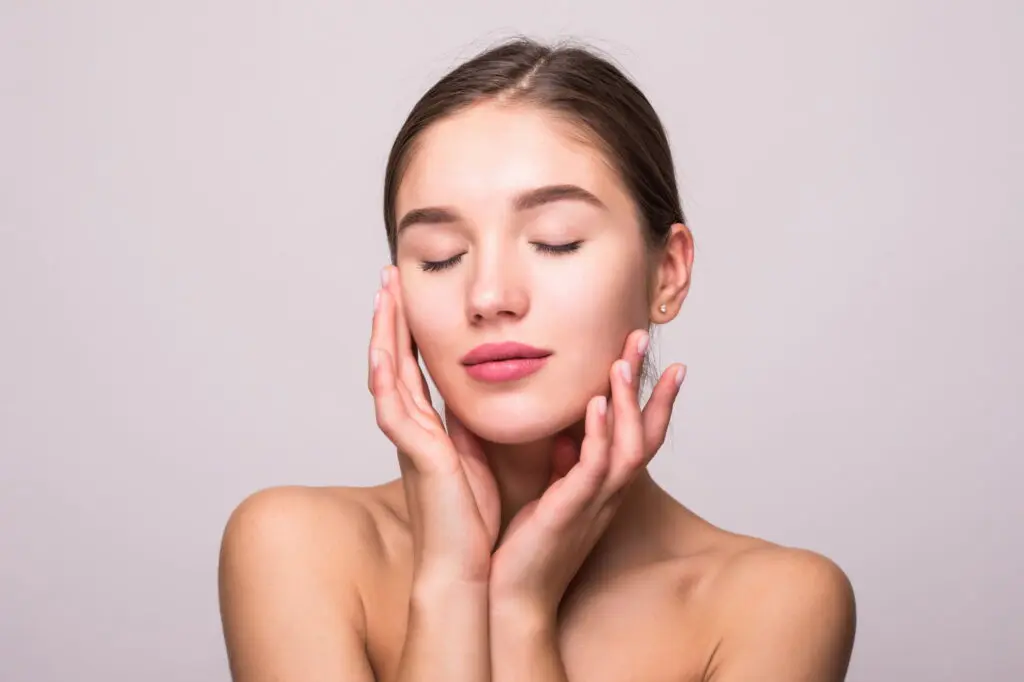 Against a plain light background, a young woman with bare shoulders gently touches her face and closes her eyes, reflecting on the refreshing effects of a hydrafacial at Refresh MedSpa in Cape Coral, FL.